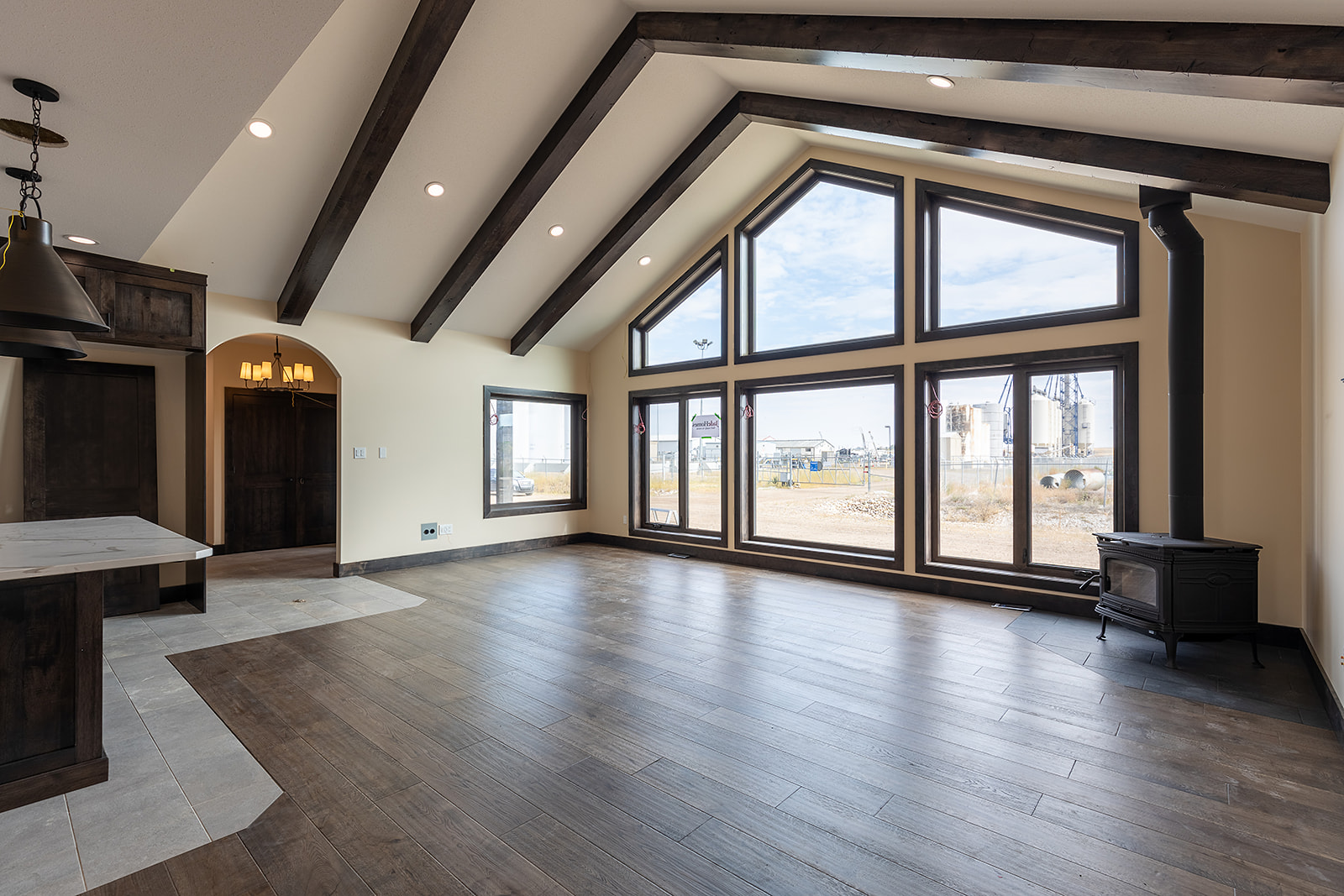 Spacious, sunlit room with large triangle-top windows, dark wood beams, and hardwood floors. A stove is in one corner, creating a cozy atmosphere.