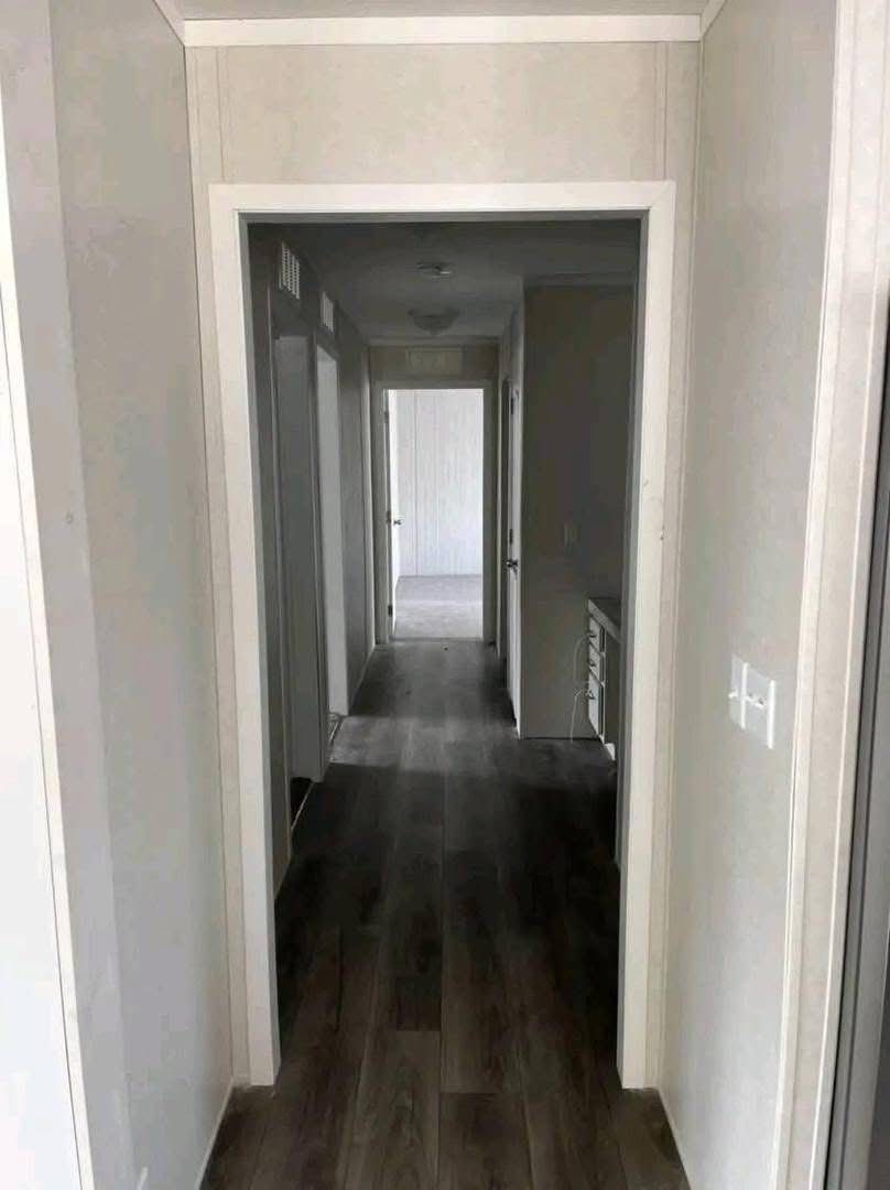 Narrow hallway with light gray walls and dark wood flooring leading to a distant open door at the end. The space appears clean and minimalistic.