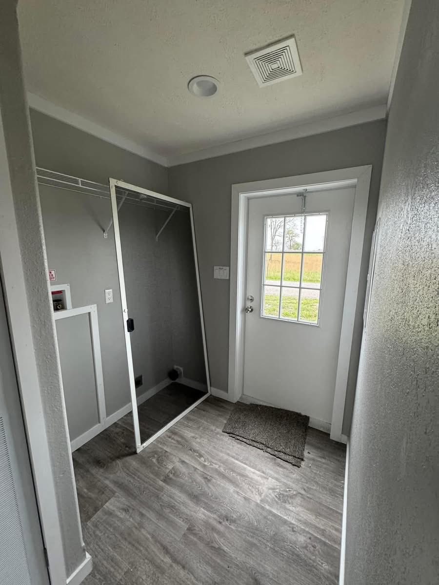 A small, gray-walled entryway with wooden floors and a glass-paneled door. A leaning mirror and doormat add a simple, functional touch.