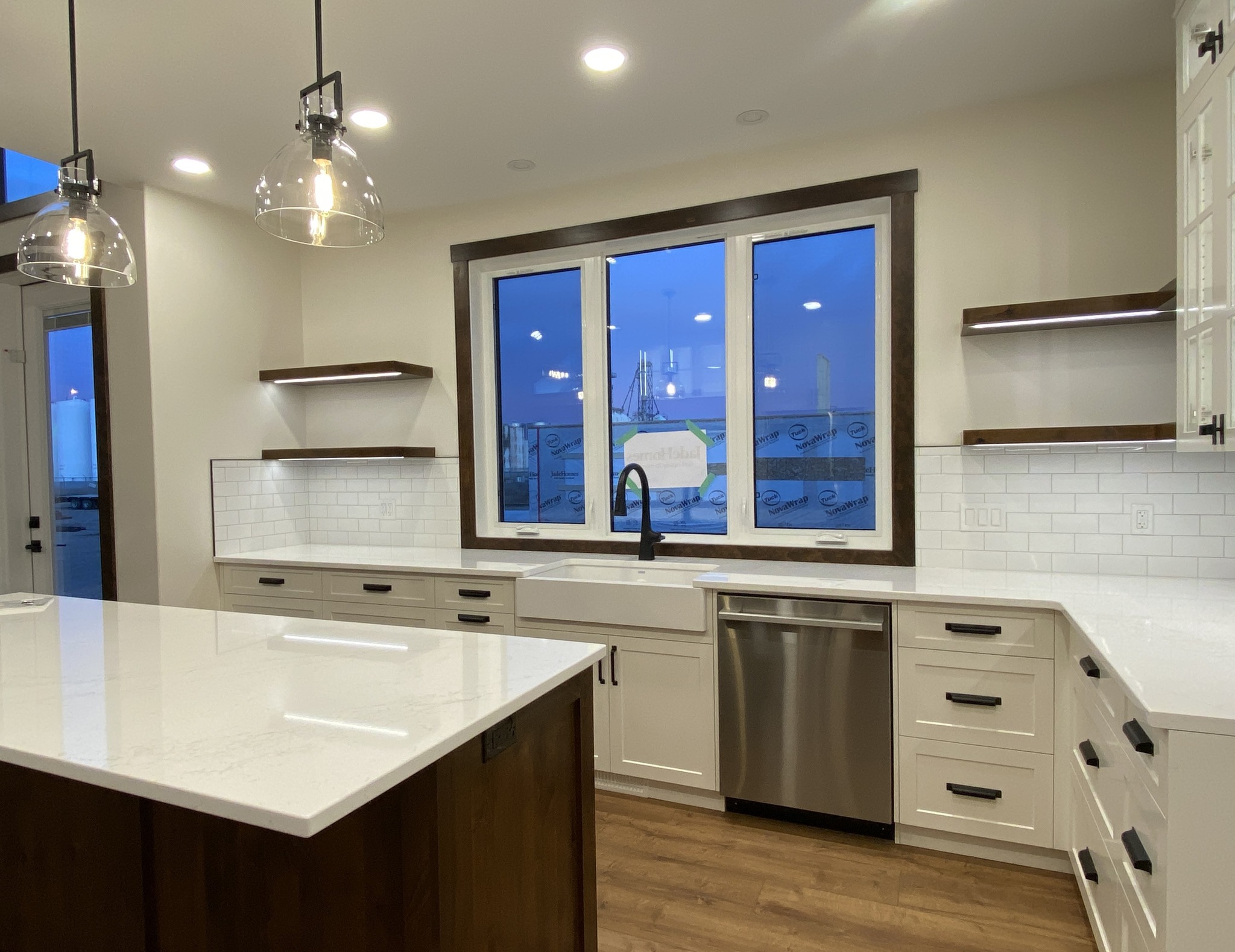 Modern kitchen with white cabinets, a large island, and stainless steel appliances. Pendant lights hang above, and a window shows a twilight view.