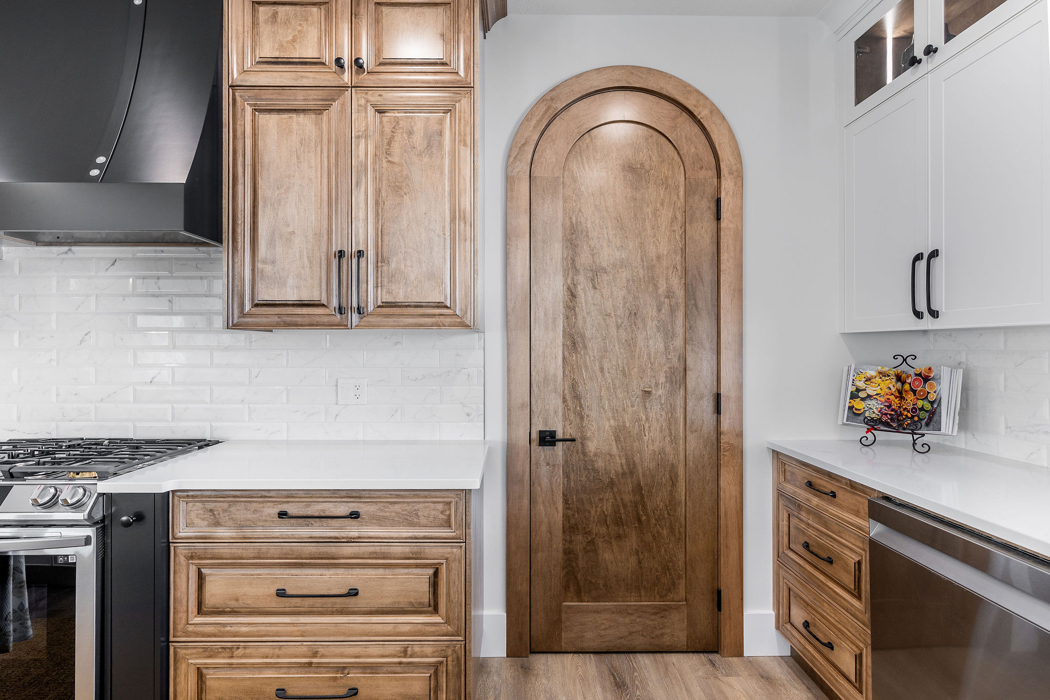 A kitchen with wooden cabinets, a stainless steel stove, white countertops, brick-style backsplash, and an arched wooden door. A floral decor piece adds warmth.