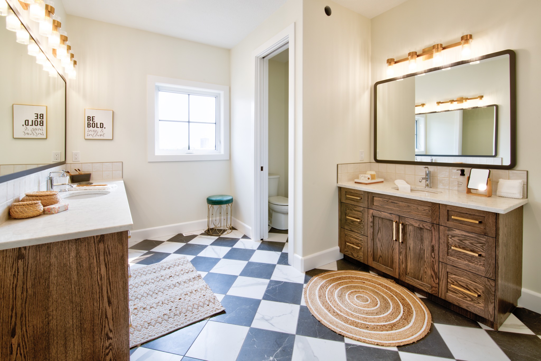 Spacious bathroom with checkered black-and-white floor, two wooden vanities, bright lighting, circular rug, and motivational wall art, creating an inviting ambiance.