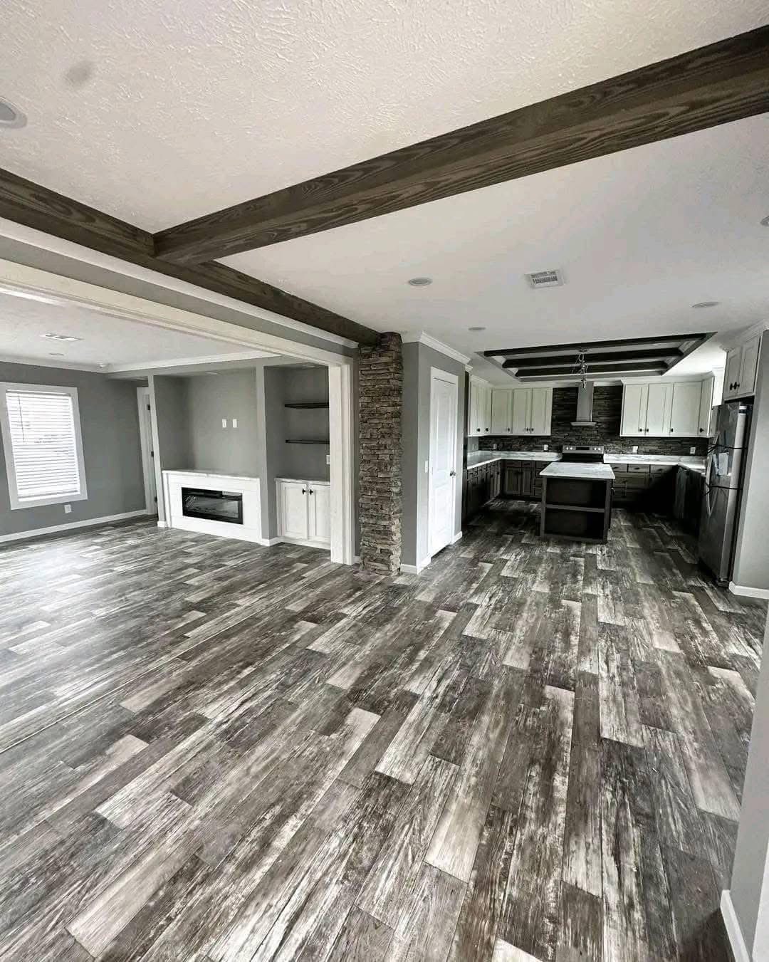 Spacious open-plan room with dark wood beam ceiling, modern kitchen, white cabinets, stone accent wall, and gray wood-look flooring. Cozy, contemporary feel.