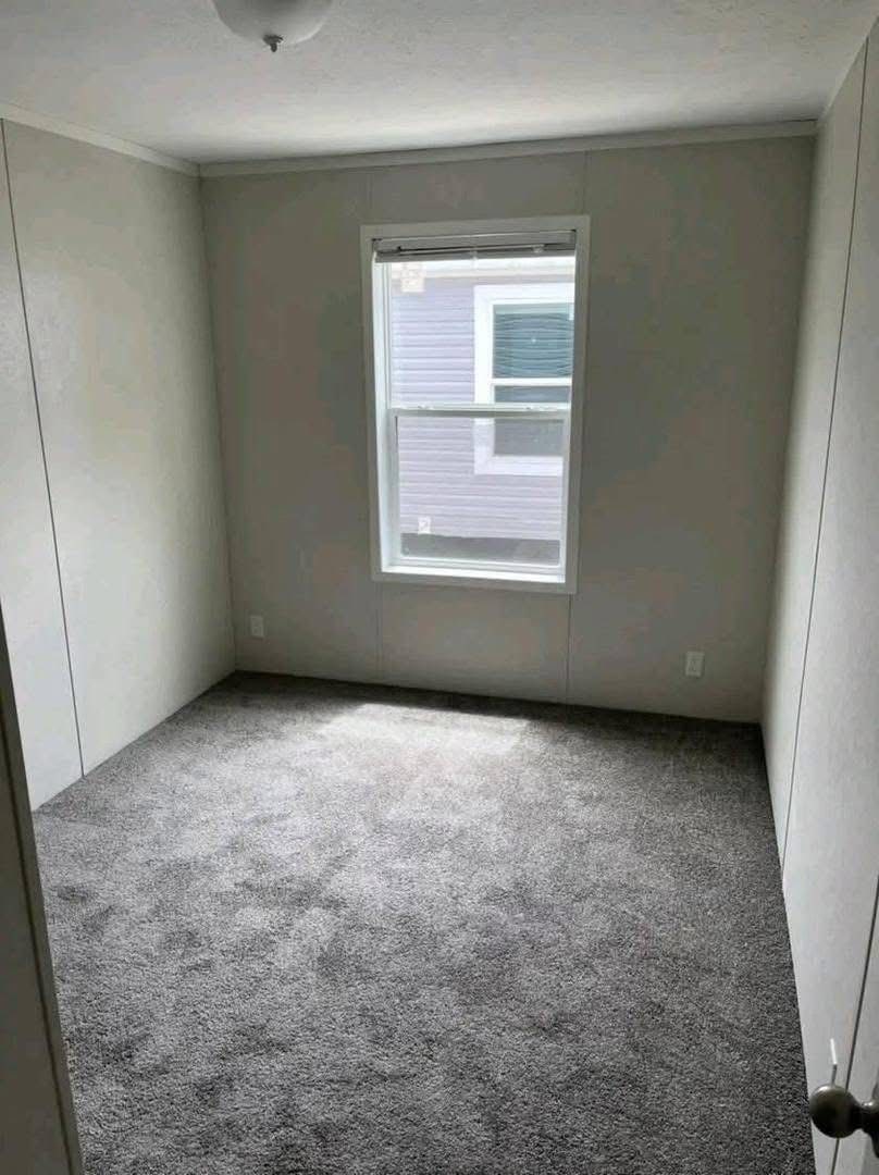 Small, empty room with light gray walls, a window revealing a neighboring house, and soft gray carpet. The space feels neutral and minimalistic.