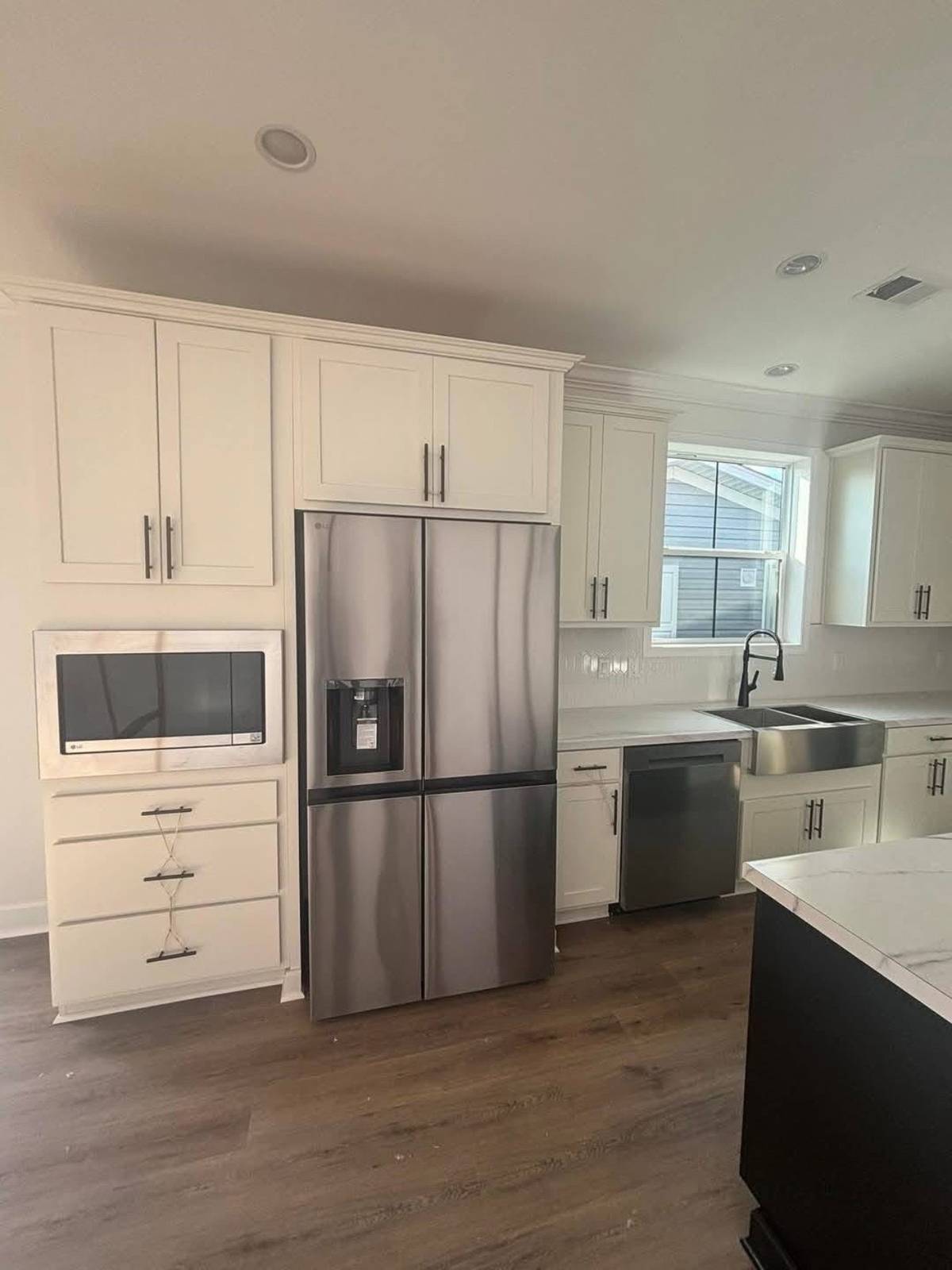 Modern kitchen with stainless steel appliances, including a fridge, microwave, and dishwasher. White cabinets, marble countertop, and wood flooring. Bright and clean.