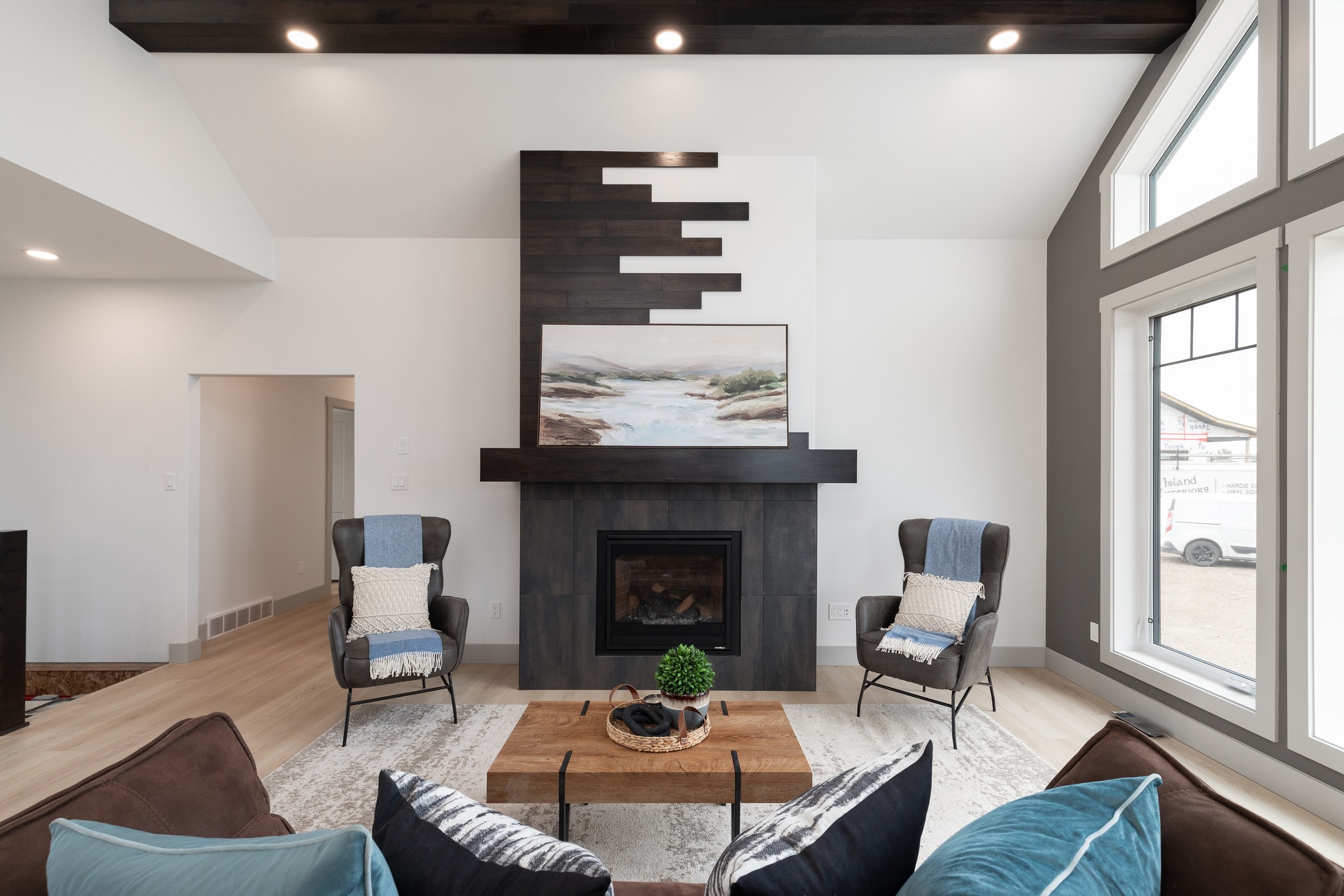 A modern bedroom with gray walls and light wood flooring. A plush gray bed is adorned with blue and white linens and a plaid throw. Three windows let in natural light.