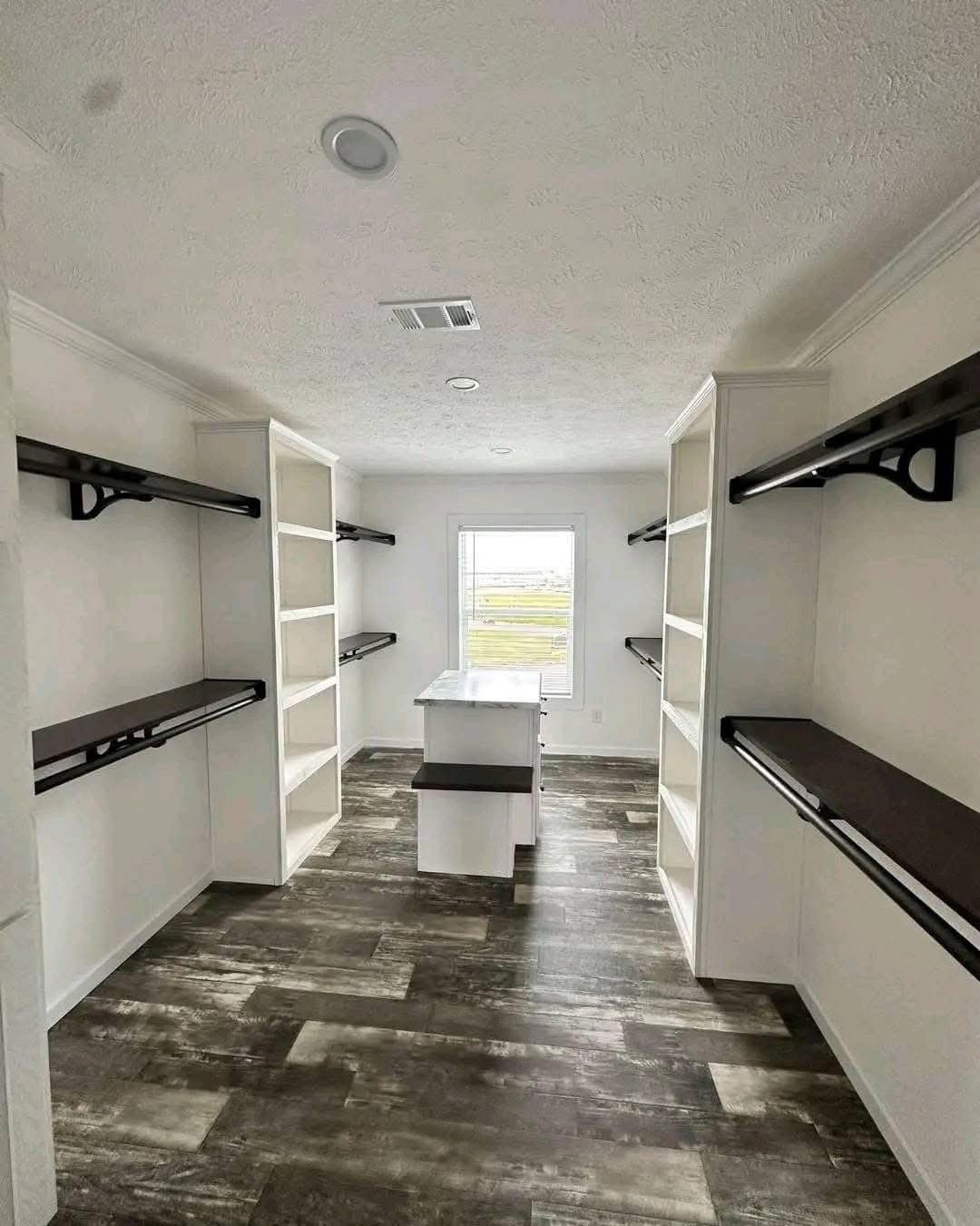 Spacious walk-in closet with white shelves and dark wooden rods on both sides. A window at the end brings natural light. Modern, organized ambiance.