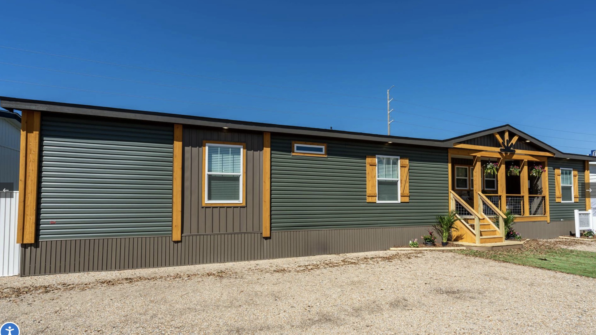 A green and brown manufactured home with contrasting wood trim is under a clear blue sky. Two windows are visible, and a small porch with steps leads to the entrance.