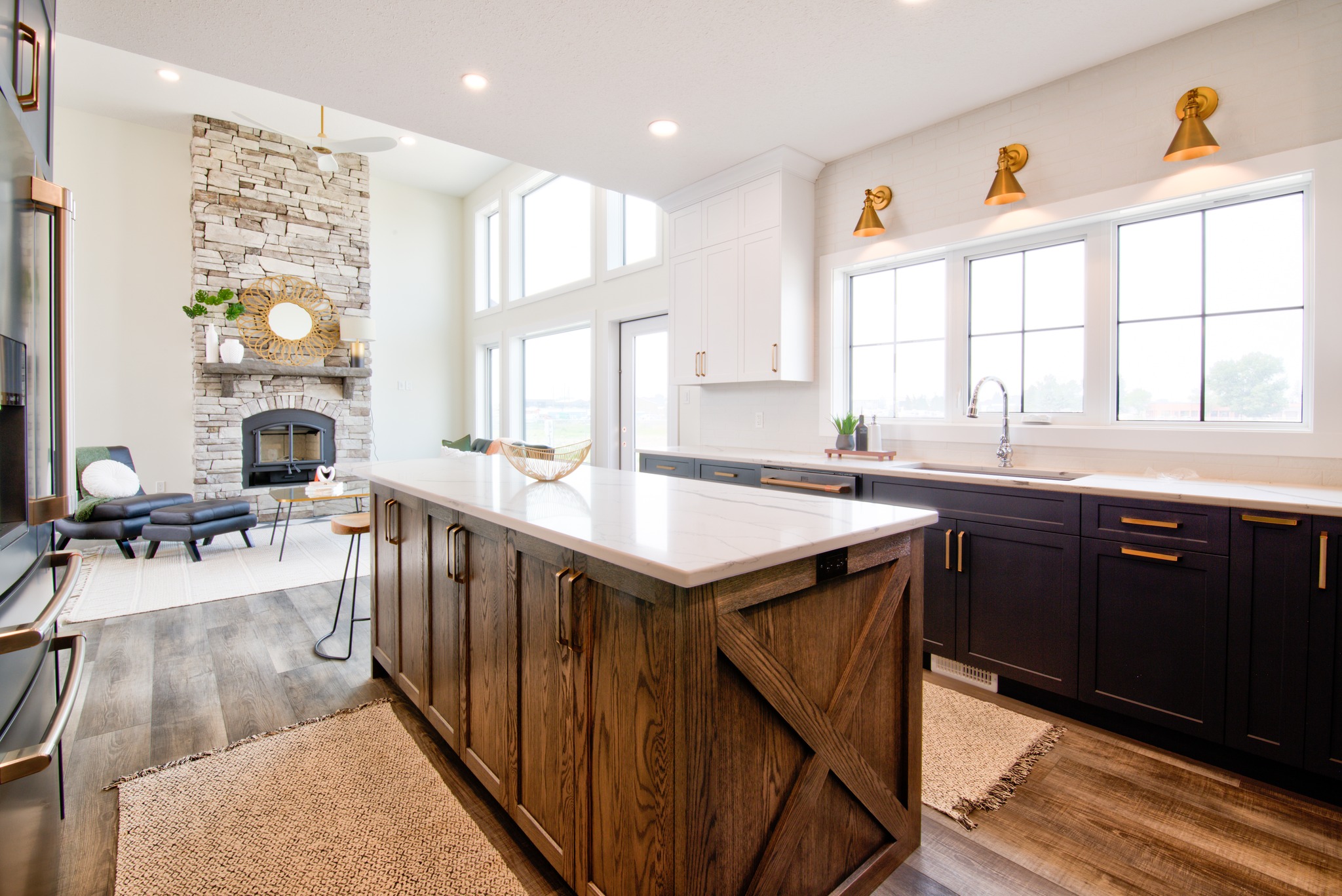 A spacious, modern kitchen with a large wooden island. Dark cabinets contrast with white countertops. Warm lighting and a stone fireplace add a cozy touch.
