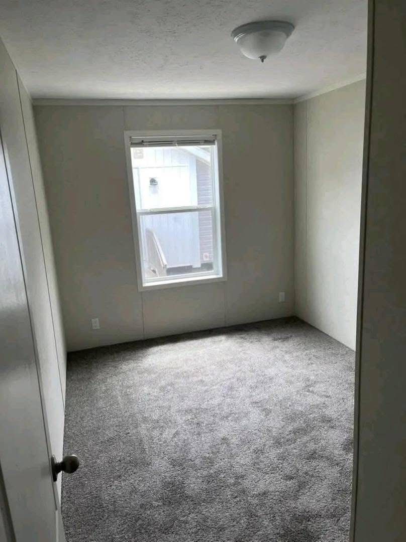 Small, empty room with light gray carpet, pale walls, and a single window letting in natural light. Ceiling light fixture in the center.