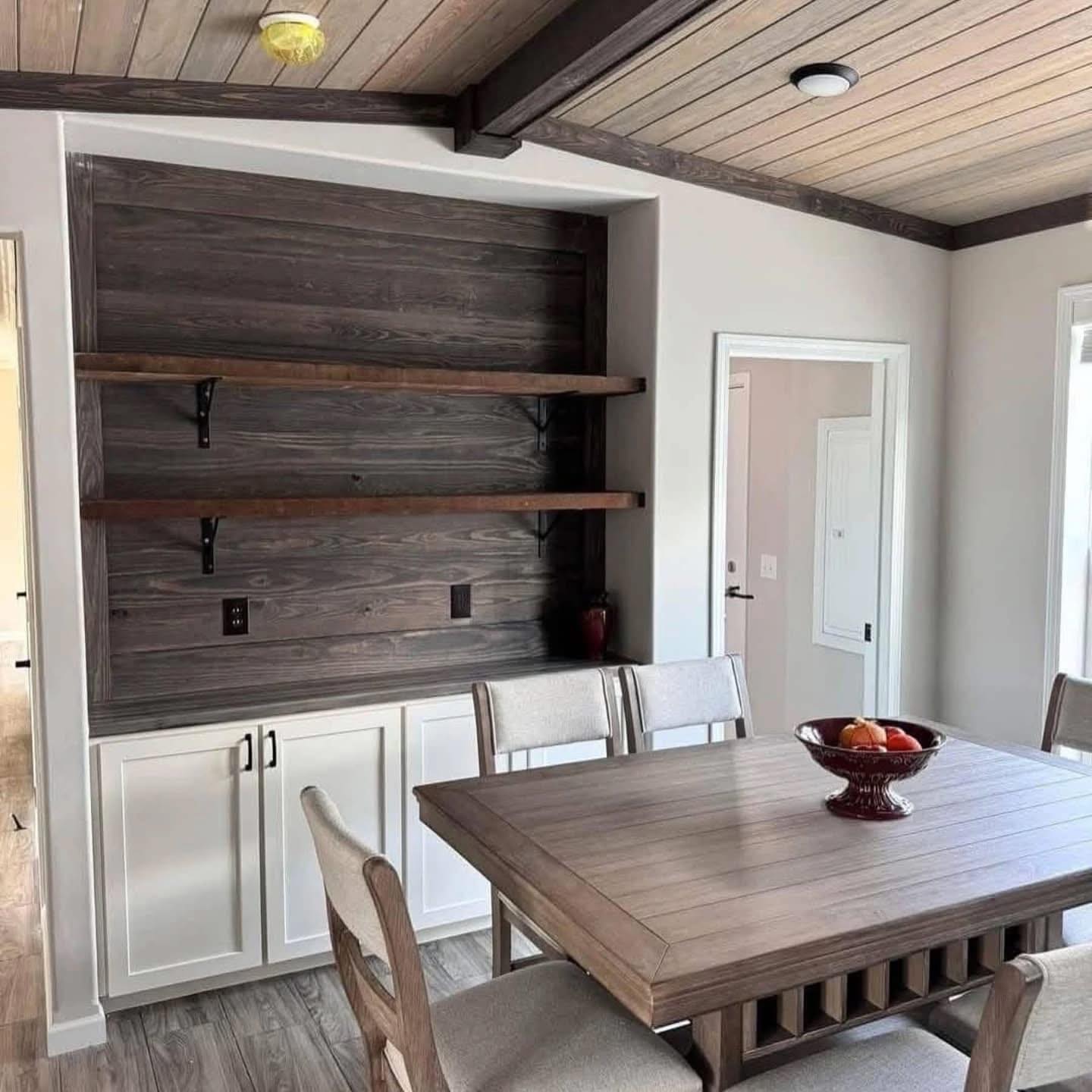 Cozy dining room with wooden table, padded chairs, and a bowl of fruit. Features rustic shelves on a dark wood accent wall with white cabinets.