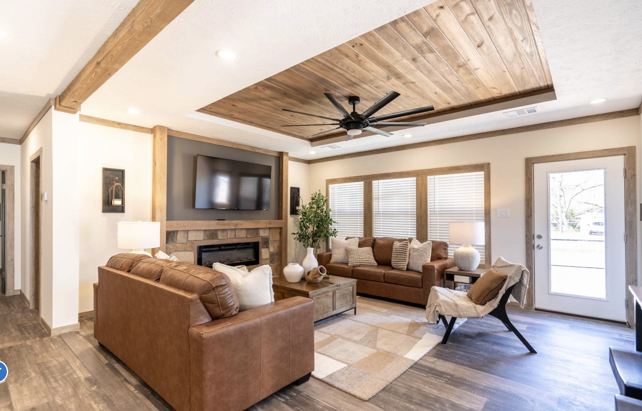 Cozy living room with brown leather sofas, wooden ceiling panels, and a large ceiling fan. A TV is mounted above the fireplace. Natural light streams in through windows and a glass door, enhancing the neutral tone decor.