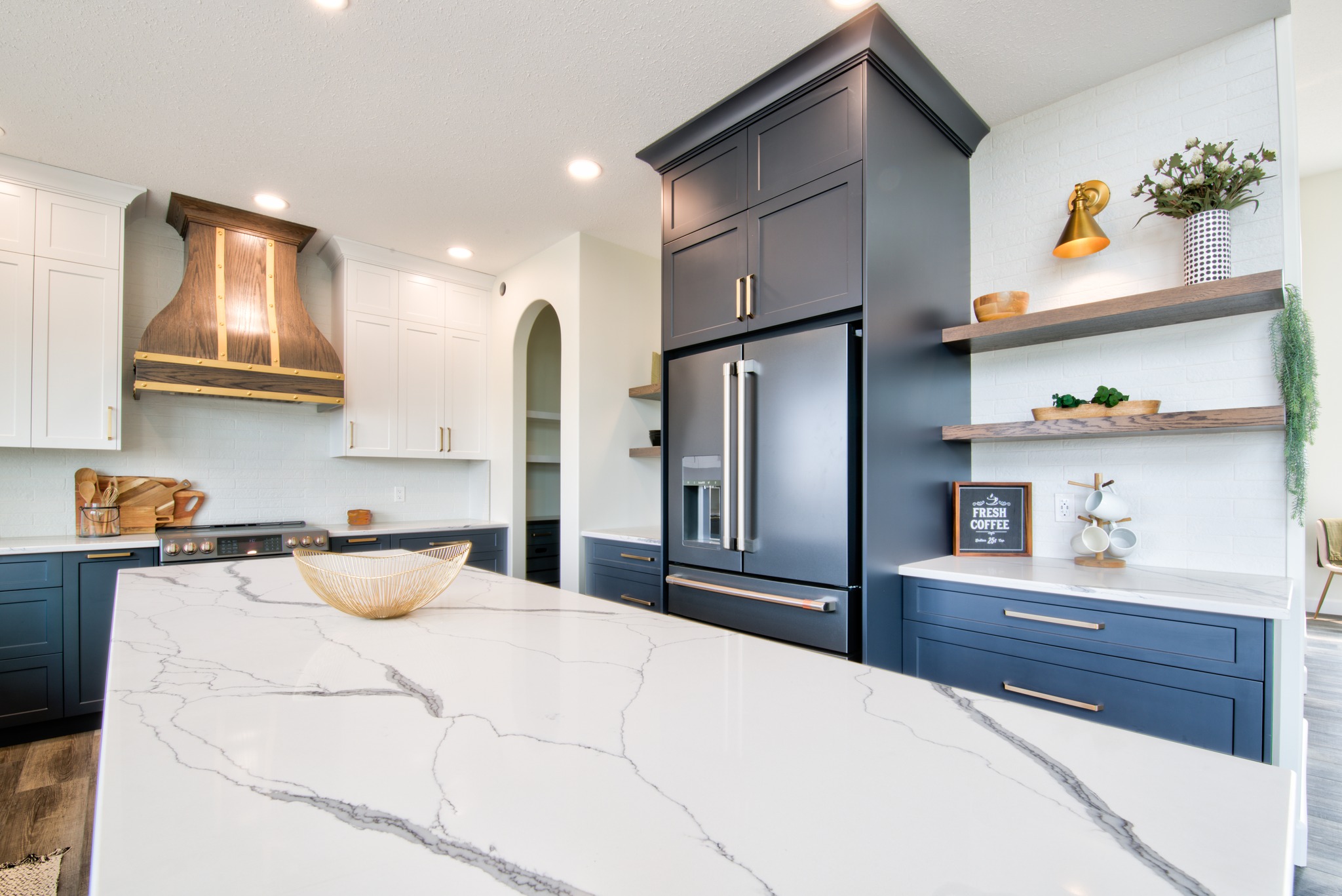 Modern kitchen with white and navy cabinets, marble island, and stainless steel fridge. Warm lighting, float shelves, and a stylish range hood.