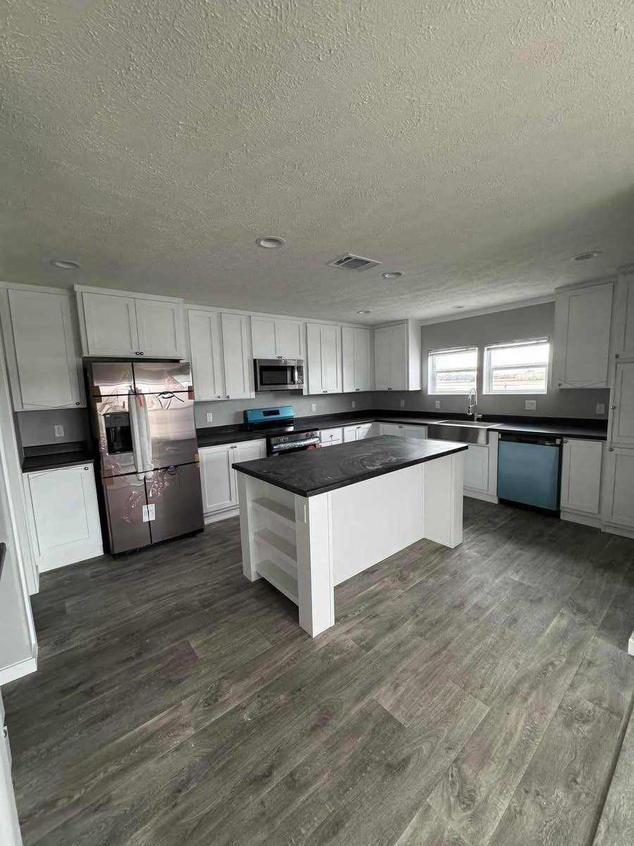 Modern kitchen with gray wood floors, white cabinets, and dark countertops. Stainless steel appliances and a central island create a sleek, spacious look.