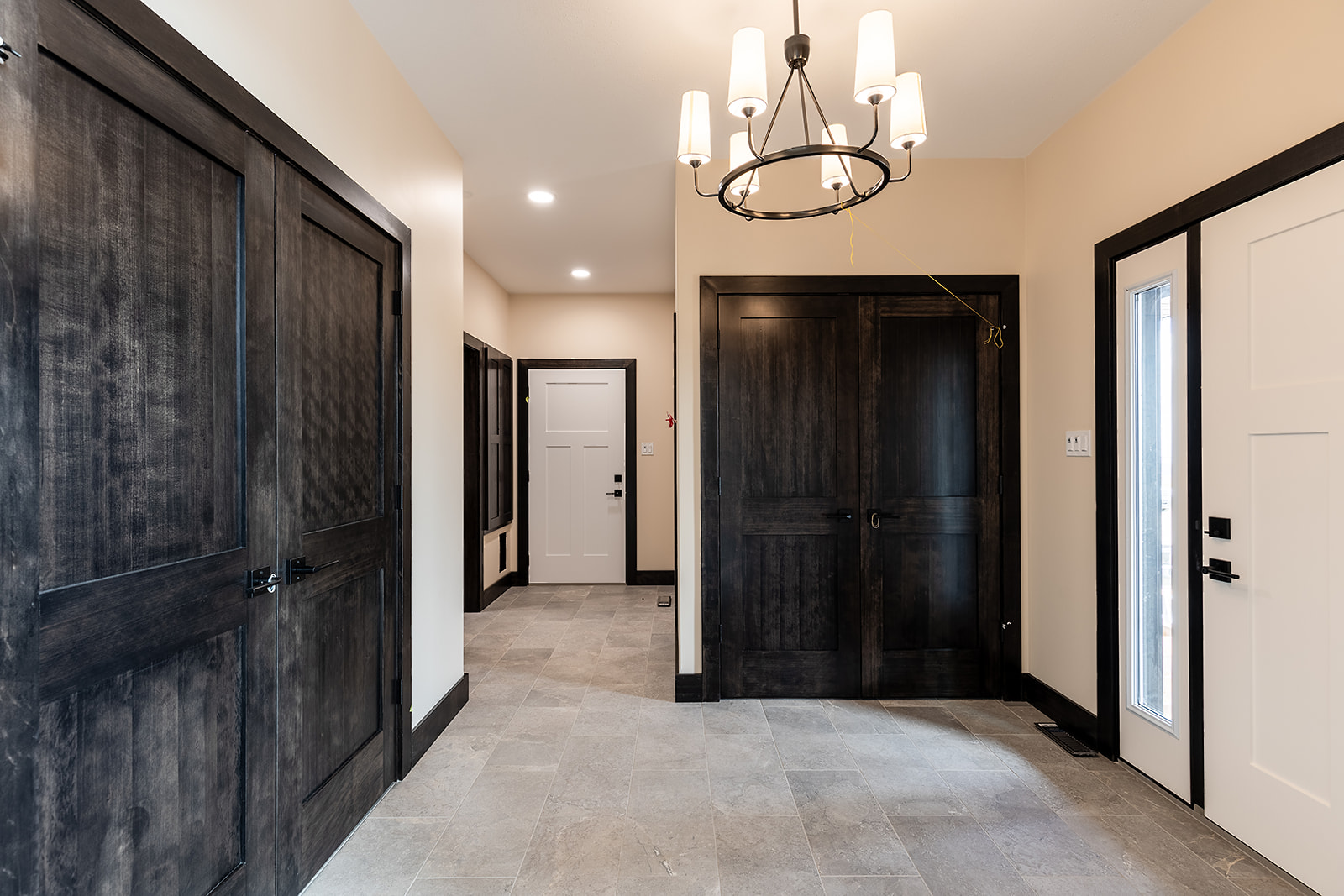 Spacious hallway with beige walls, gray tiled floor, and large dark wooden doors. A modern chandelier hangs from the ceiling, creating a welcoming atmosphere.