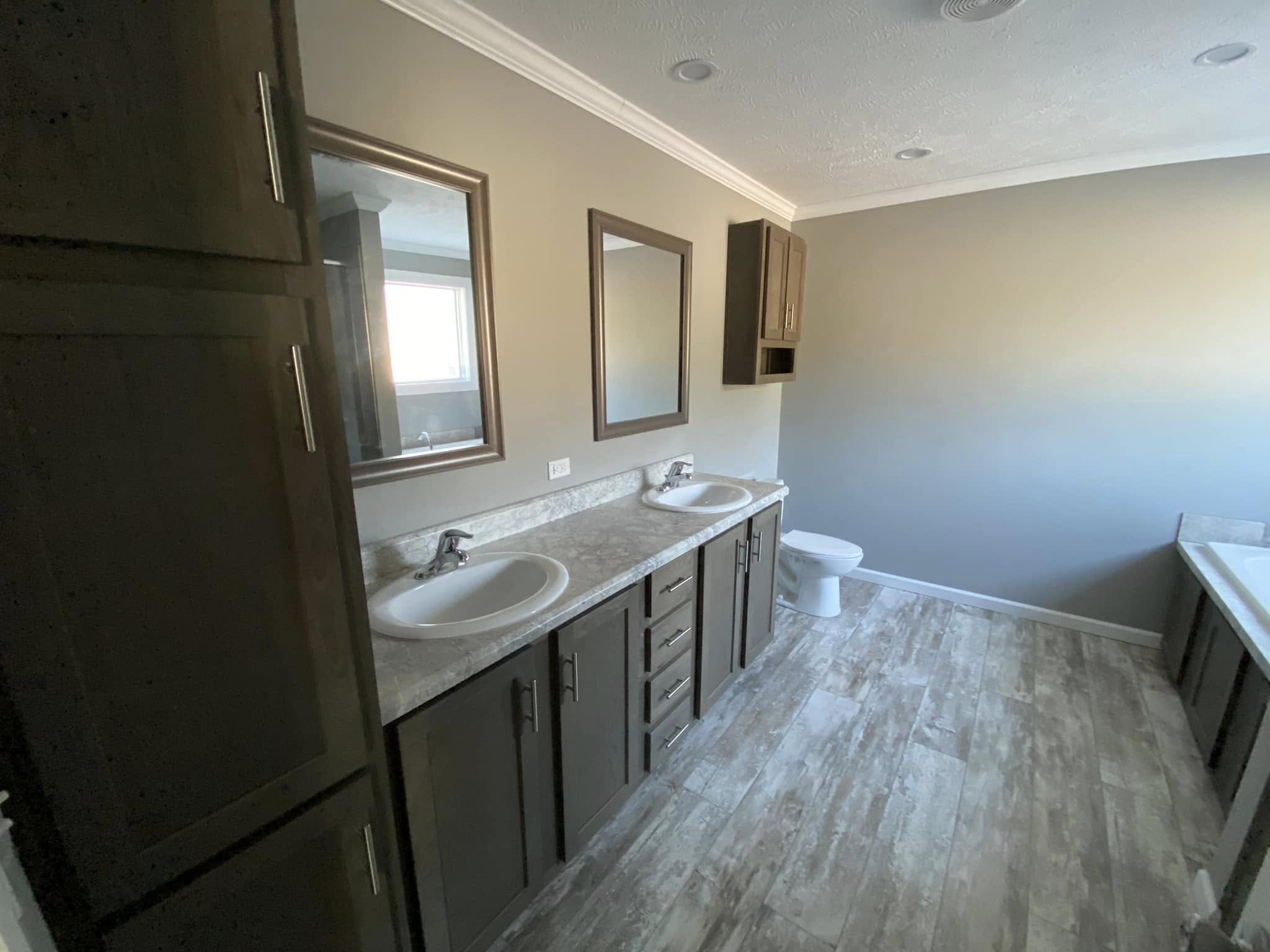 Spacious bathroom with dual sinks on a marble countertop, two mirrors, wooden cabinets, and a bathtub. Light gray tiles add a modern touch.