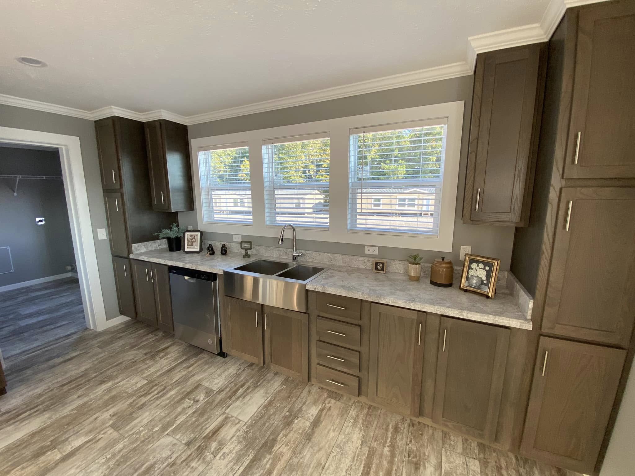 Modern kitchen with brown cabinets, stone countertops, and a stainless steel sink under three large windows. Wooden floor adds a warm touch.