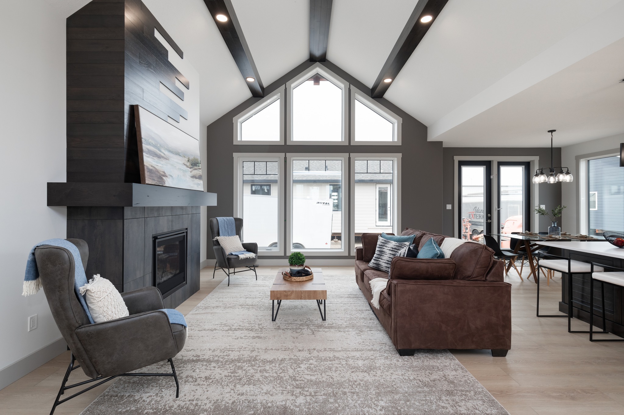 Spacious modern living room with high ceilings, large windows, and a cozy fireplace. Brown sofa and chairs create a warm, inviting atmosphere.