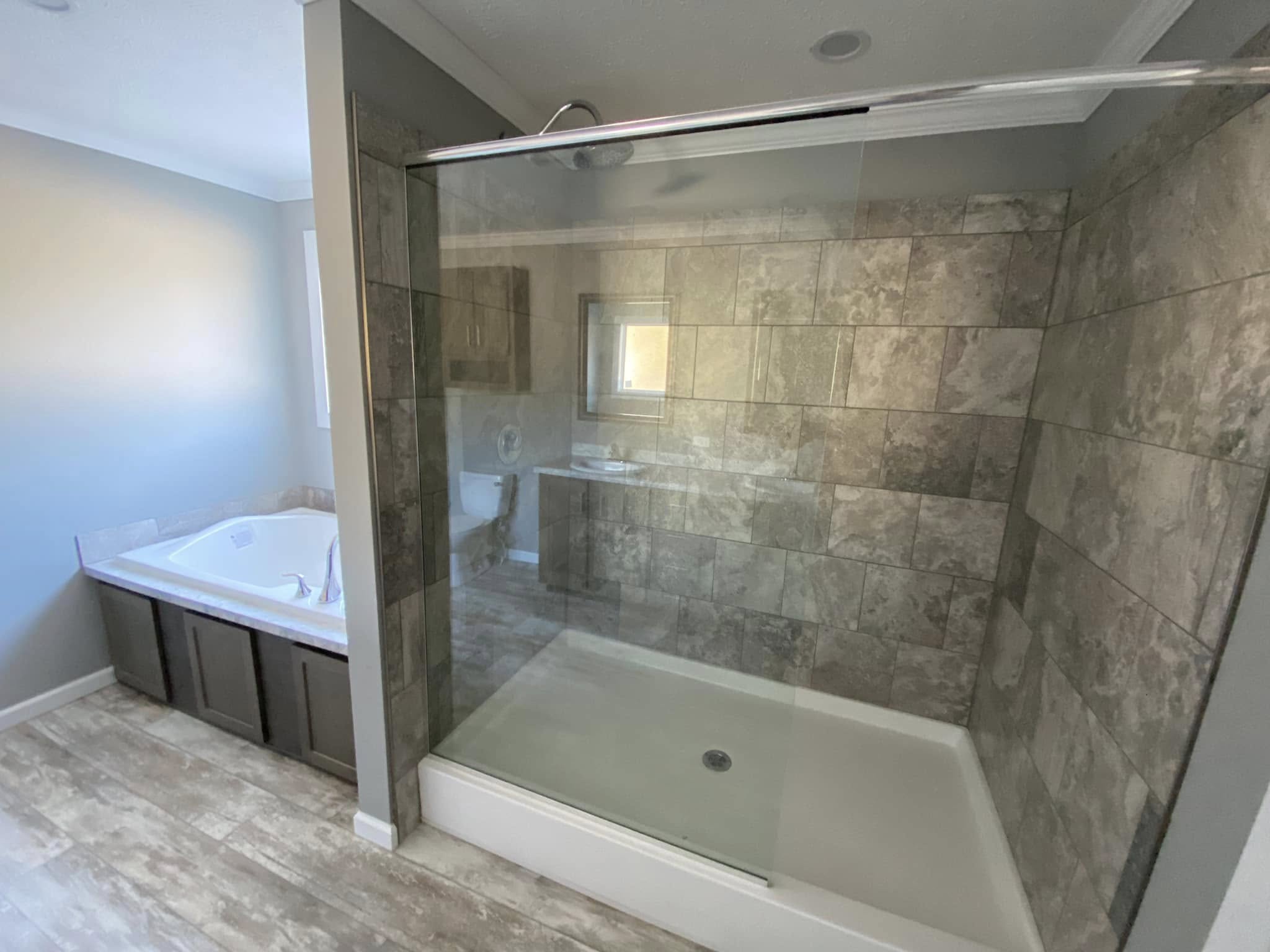 Modern bathroom with a tiled walk-in shower featuring glass doors. Adjacent is a built-in bathtub with a wood-paneled base. Warm, neutral tones.