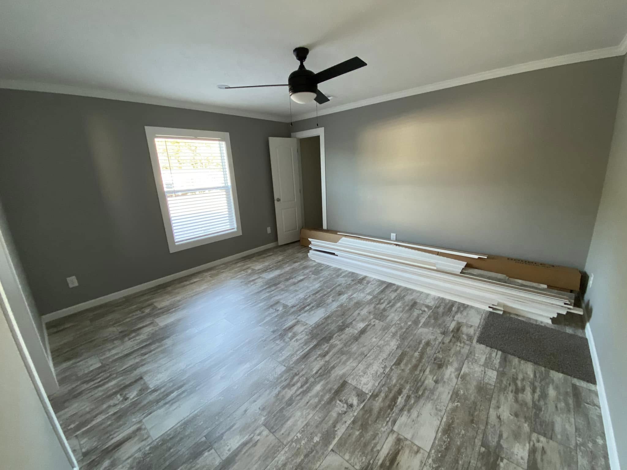 Empty room with gray walls, wooden flooring, and a ceiling fan. A window with blinds lets in light. White trim pieces are stacked against the wall.