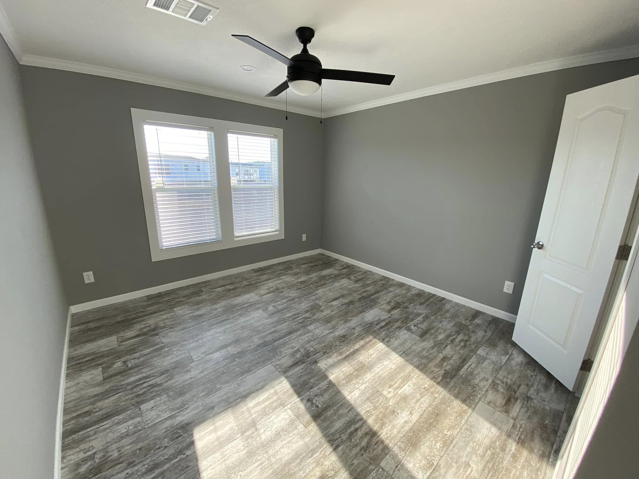 A minimalist empty room with gray walls, wooden floor, and two windows. A ceiling fan is centered above. Sunlight casts shadows on the floor.