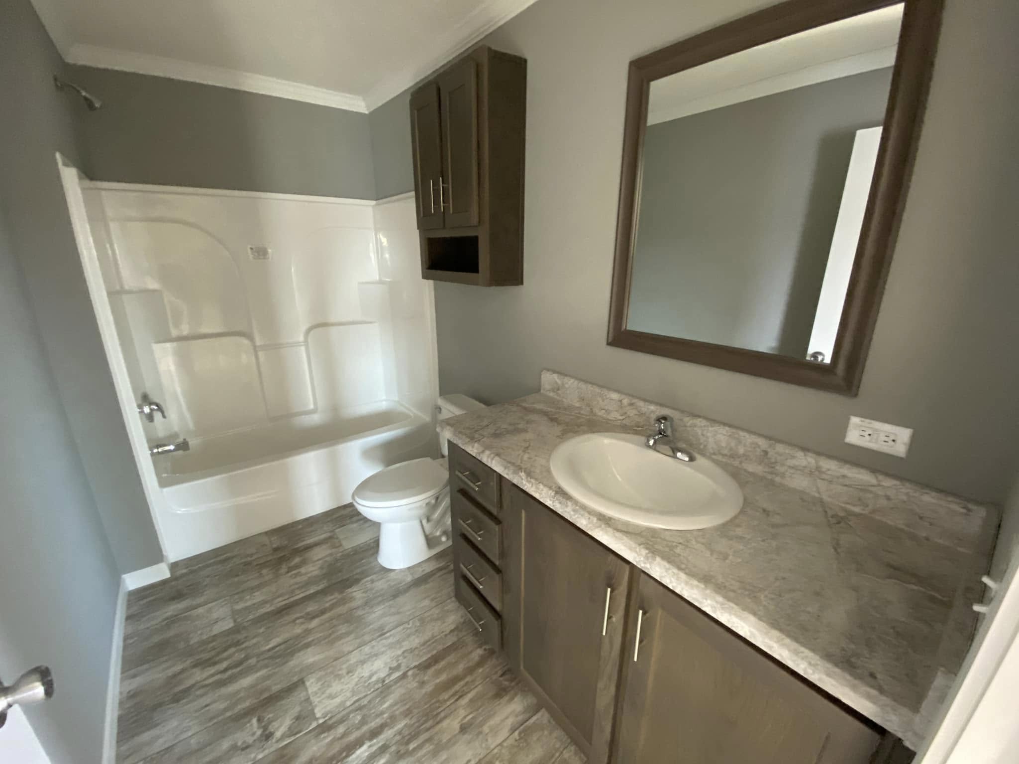 A modern bathroom with gray walls, wood-look flooring, a dark wood vanity with a marble countertop, a large mirror, and a white bathtub-shower combo.
