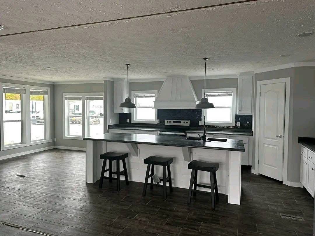 Spacious kitchen with dark wooden floors, a central island with three stools, pendant lights, white cabinets, and large windows, conveying a modern feel.