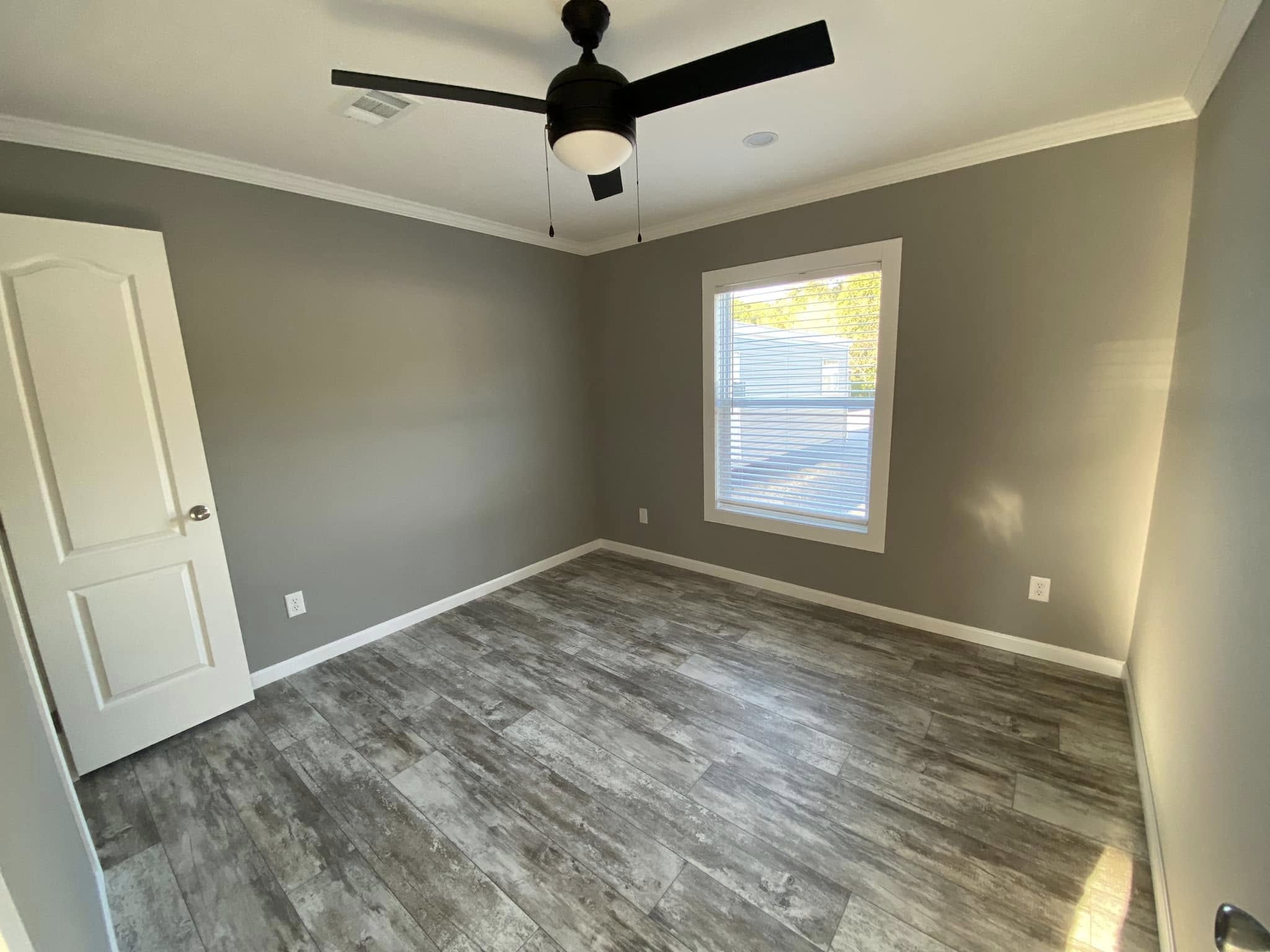 A small, empty room with gray walls, wood-look flooring, a white door, and a large window. A black ceiling fan is centered on the ceiling.