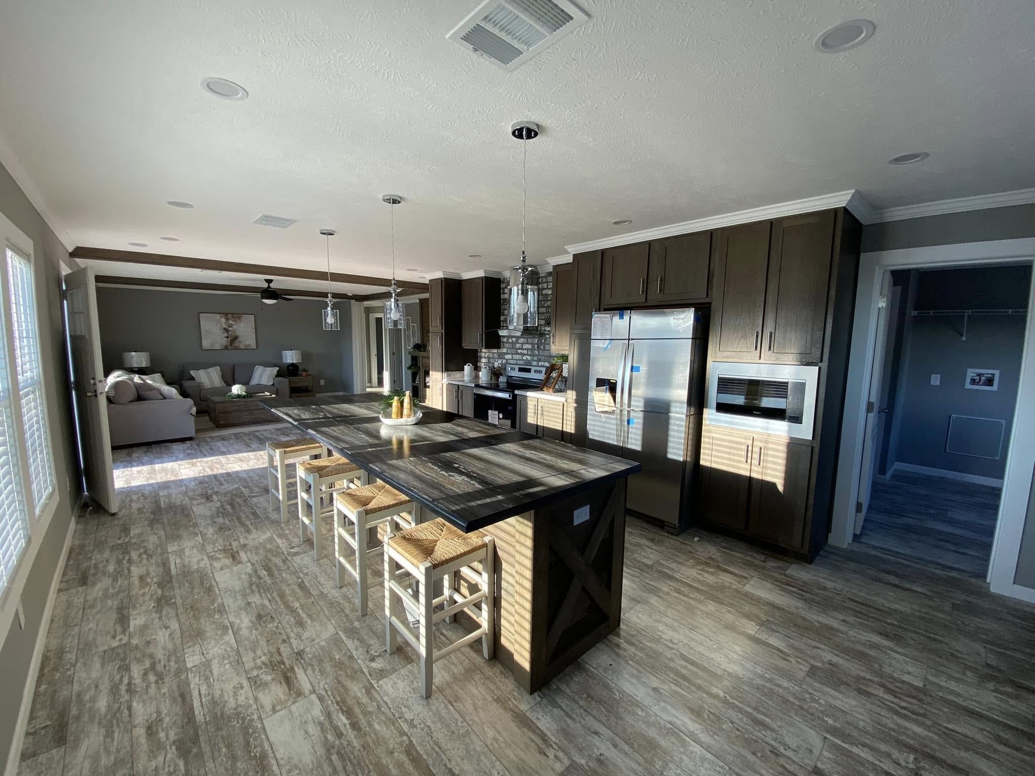 Modern kitchen with a large island, wooden stools, stainless steel appliances, and dark cabinetry. Sunlight streams through a window, illuminating the space.