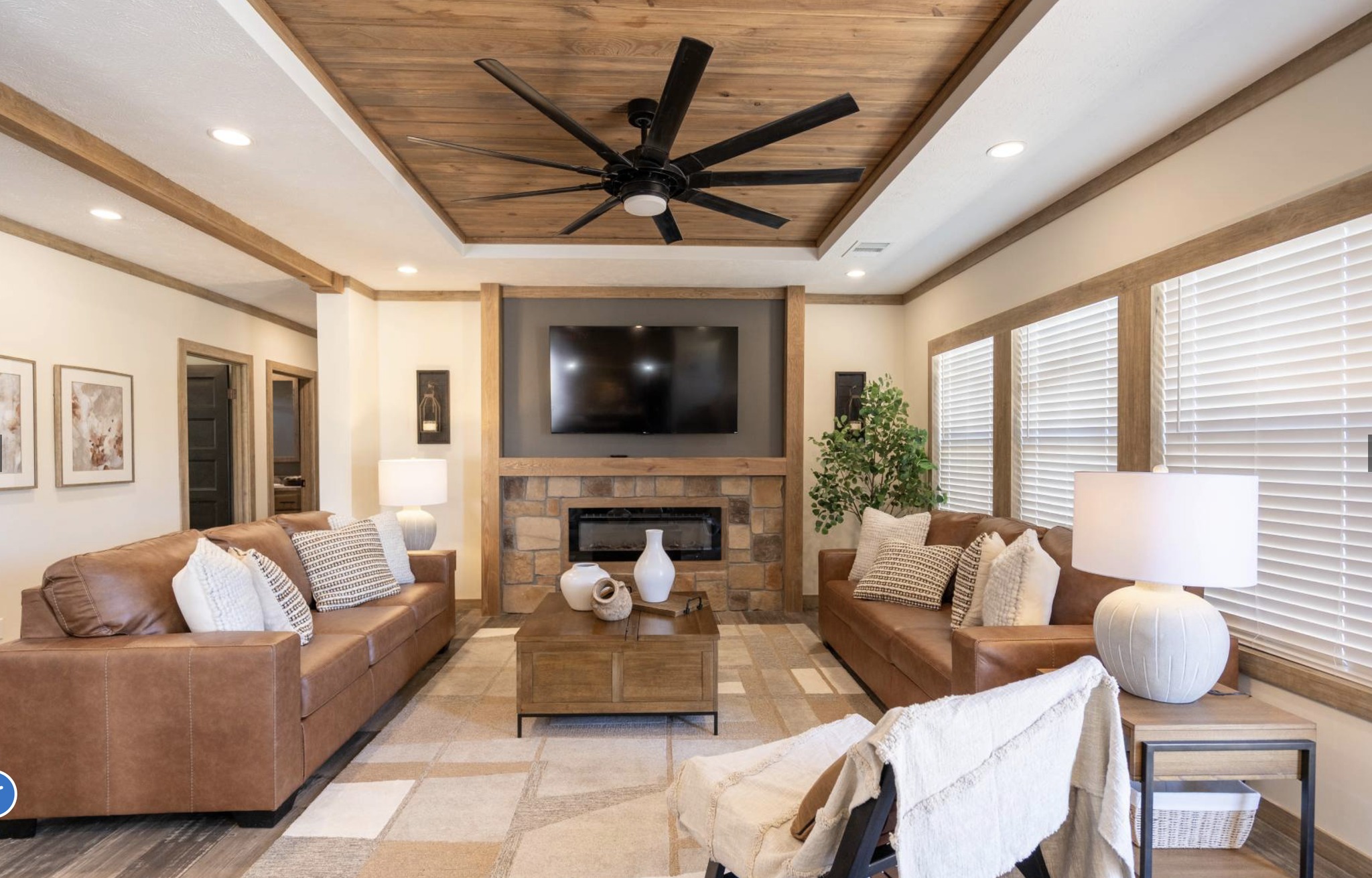 Warm, inviting living room with two brown leather sofas, a wooden coffee table, and a TV above a stone fireplace. Soft lighting and neutral tones.