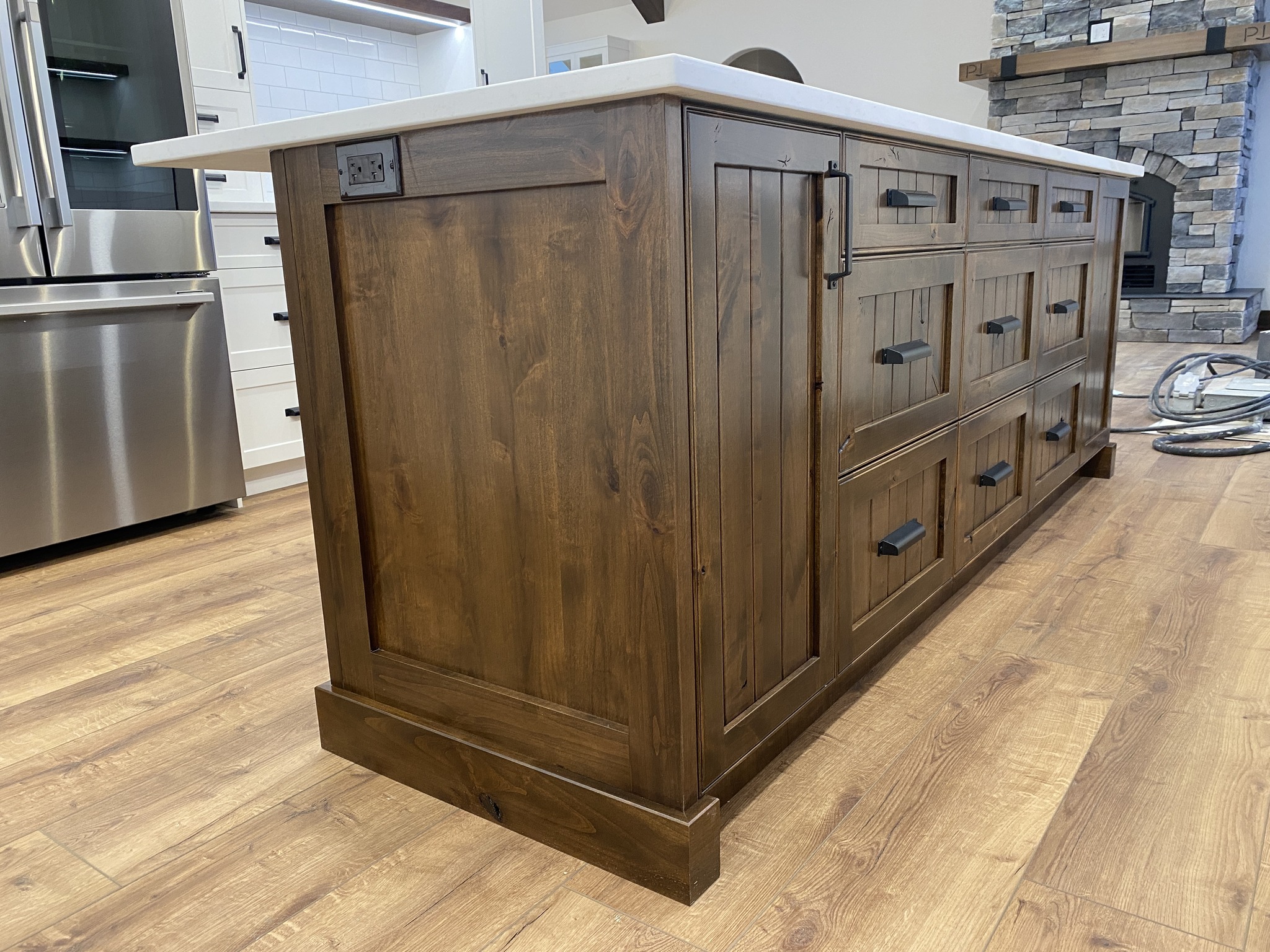 Rustic wood kitchen island with black handles and a white countertop. Surrounded by wood flooring, stainless steel fridge, and a stone fireplace.