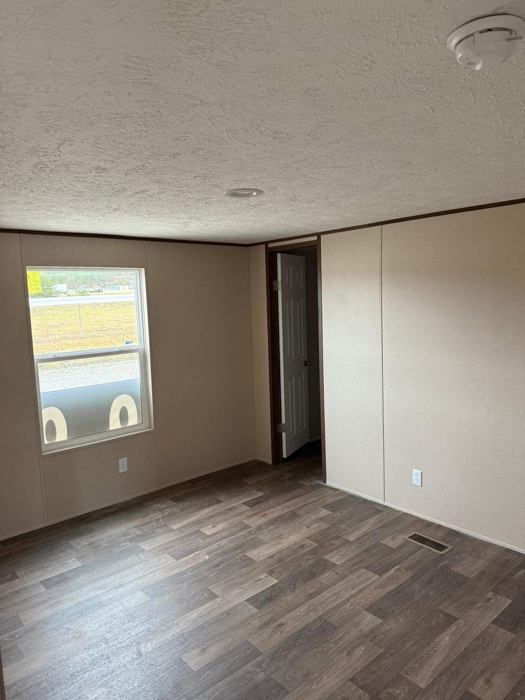 Empty room with beige walls and dark wood flooring. A single window shows a glimpse of an outside area. A partially open door leads to an adjacent room.