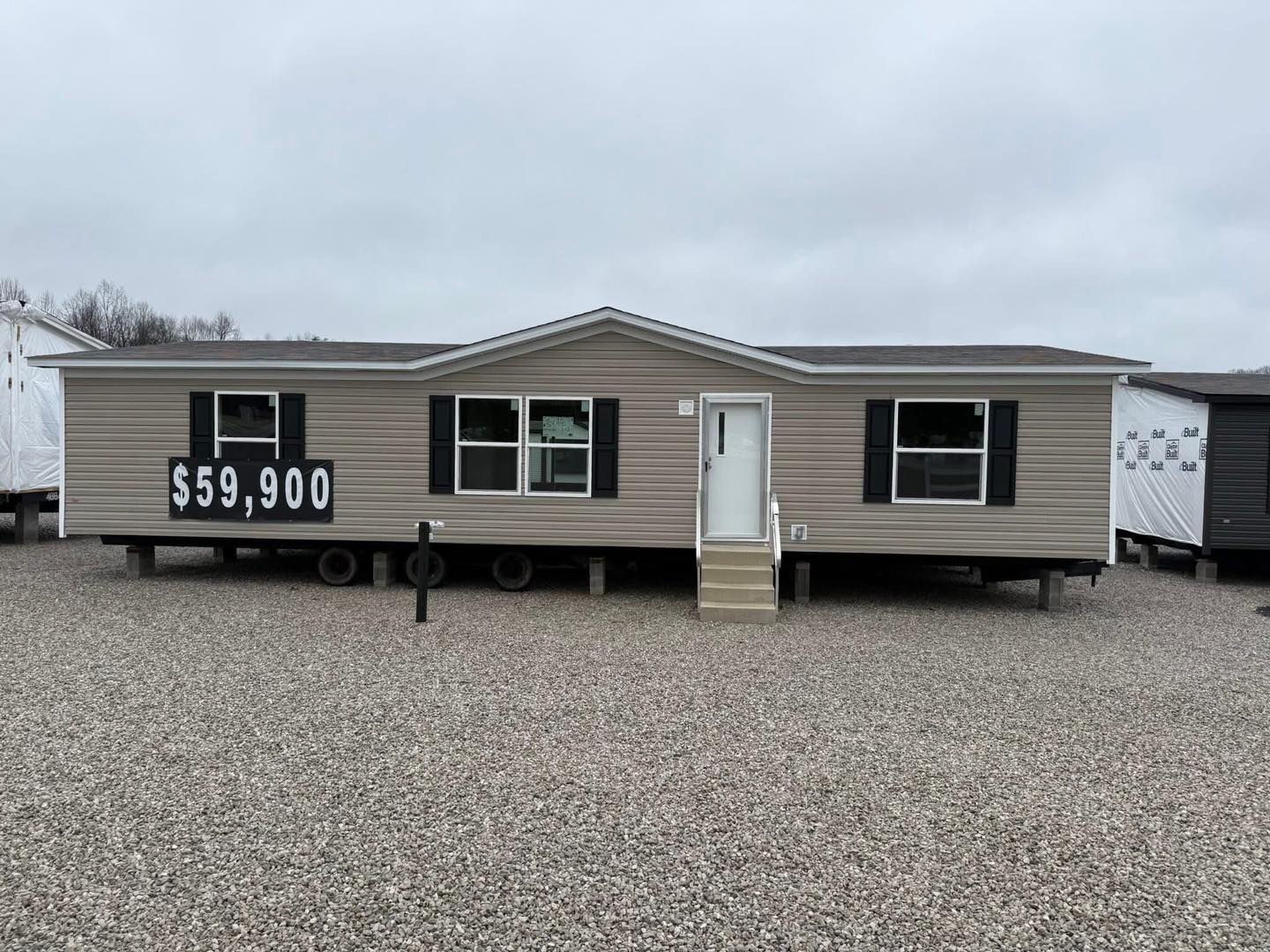 A beige manufactured home sits on gravel with a large $59,900 price sign. It features white trim, black shutters, and an overcast sky above.