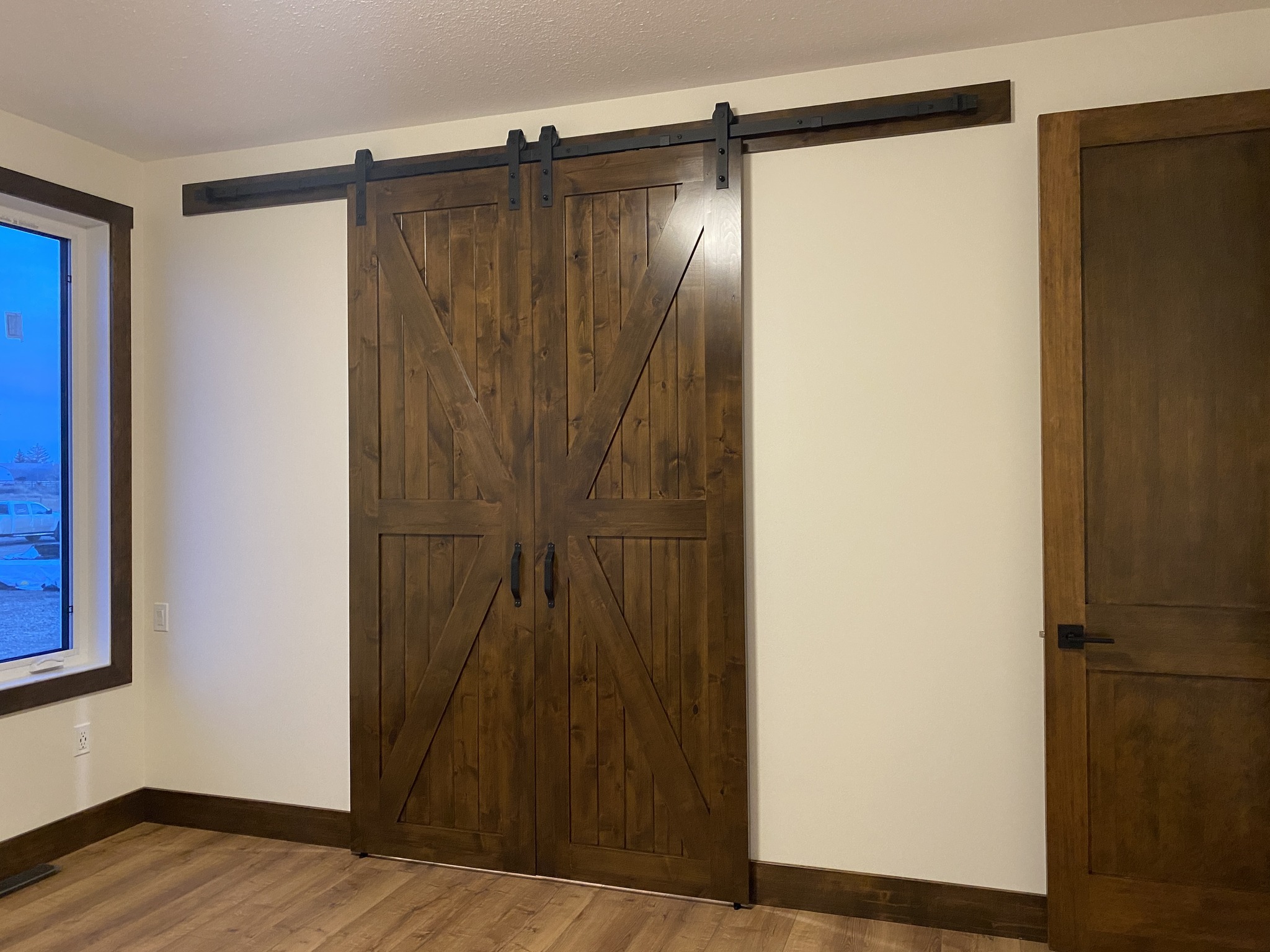 Wooden sliding barn doors on a black track in a room with light walls and wooden floor. A window and a regular door are visible on the sides.