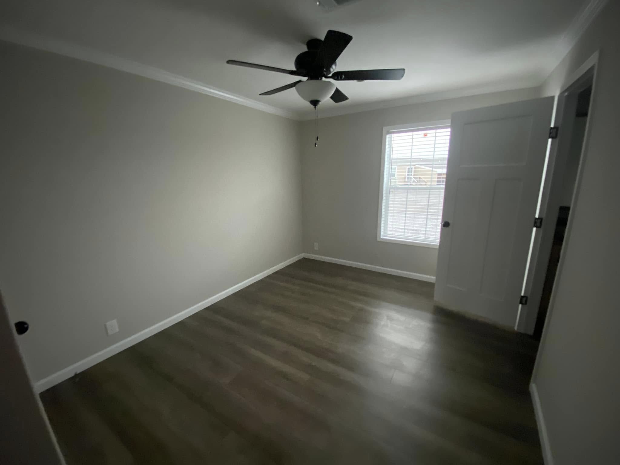 Empty room with light gray walls, dark hardwood floor, a ceiling fan, and a window with blinds. An open door suggests a quiet, minimalist space.