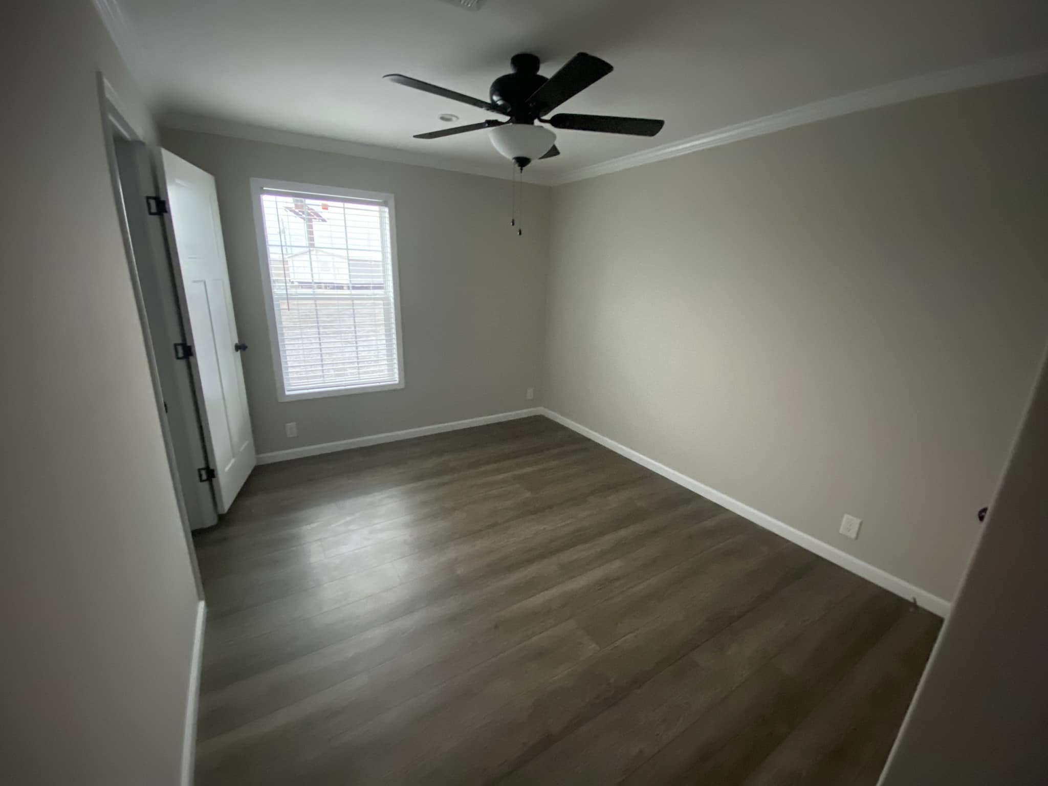 Empty room with light gray walls, dark wood floor, and a ceiling fan. A door and a window with closed blinds let in soft light, creating a calm atmosphere.