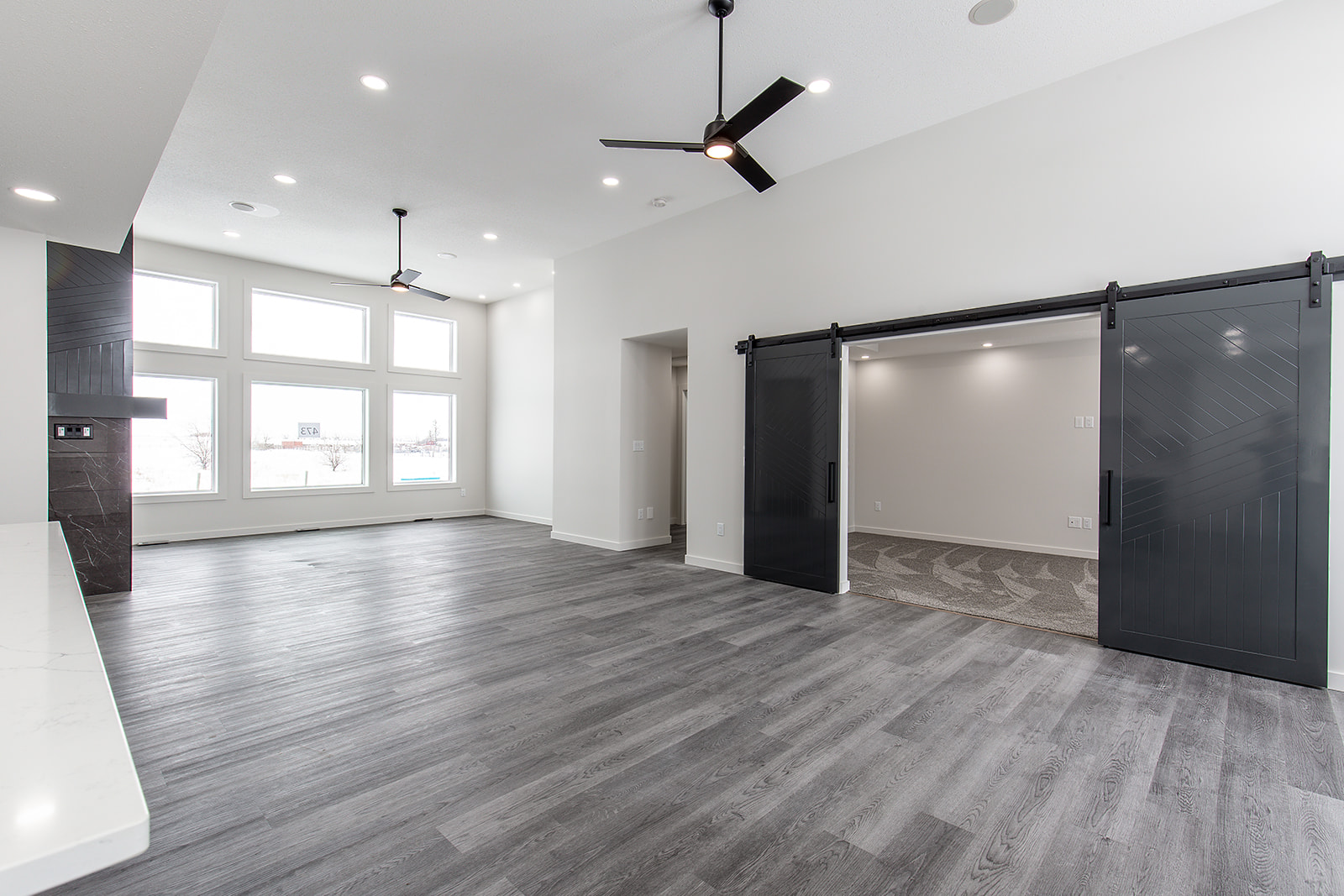 Spacious, modern living room with large windows, dark wood floors, ceiling fans, and sliding barn doors. Minimalist design, bright and welcoming.