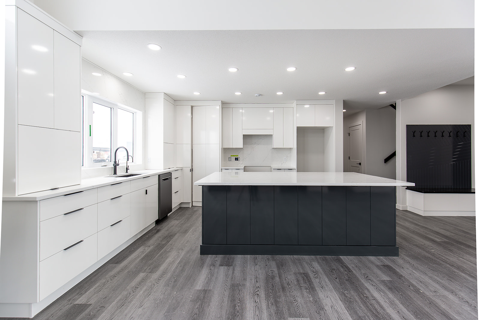 Modern kitchen with a large island, sleek white cabinets, and dark lower cabinetry. Bright space features recessed lighting and gray wood flooring.