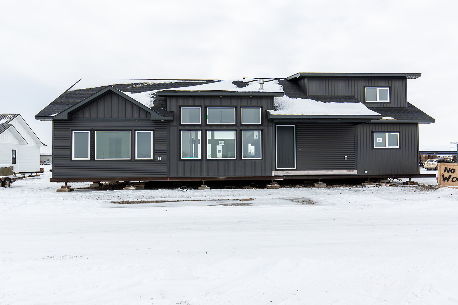 A black, two-story modular home sits on a snowy lot. The house features large windows and a modern design against a cloudy sky, conveying a cold, peaceful setting.
