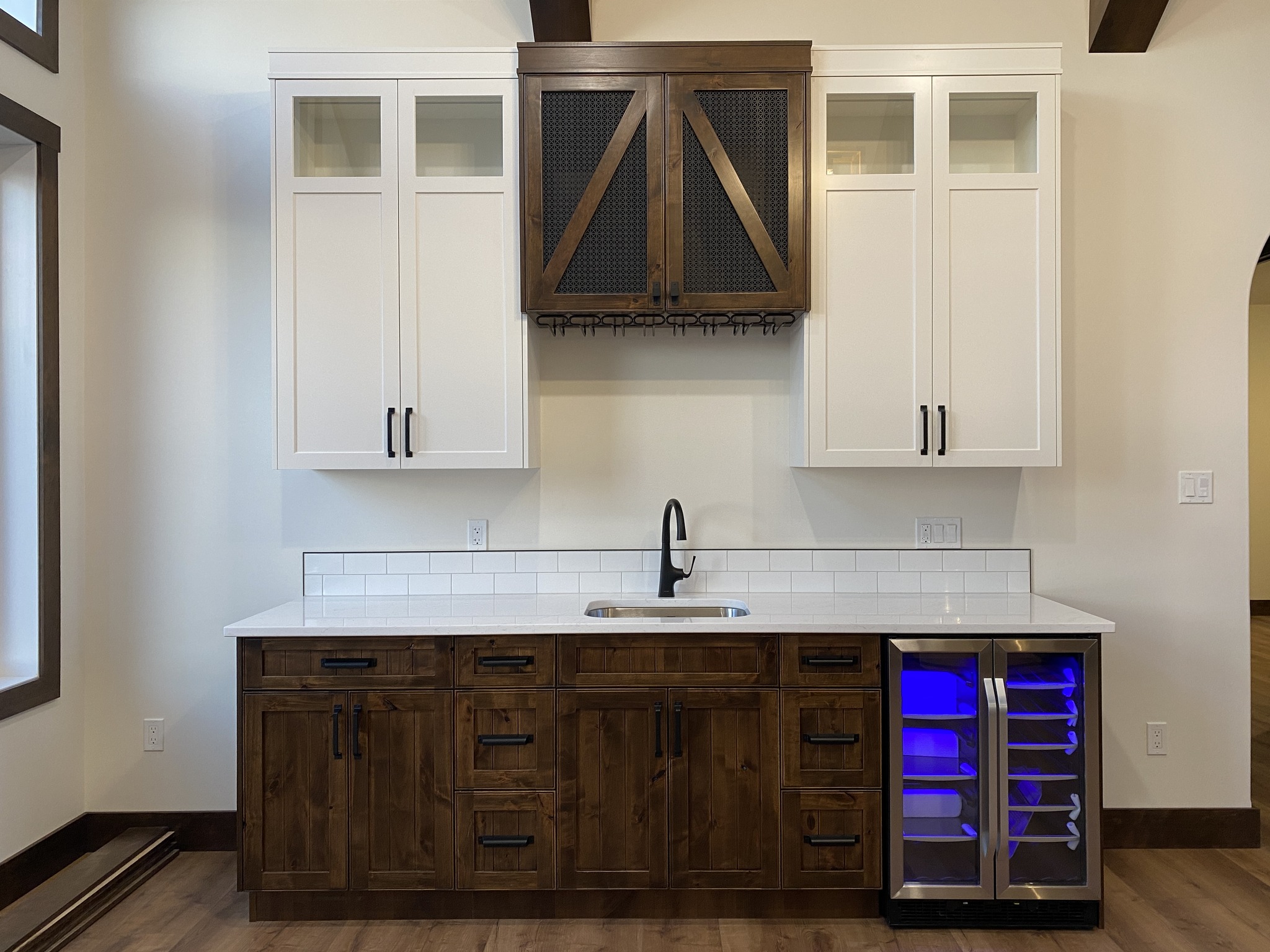 Rustic wet bar with white upper cabinets, a central farmhouse-style wooden wine rack, and a sleek mini fridge with blue lighting below.