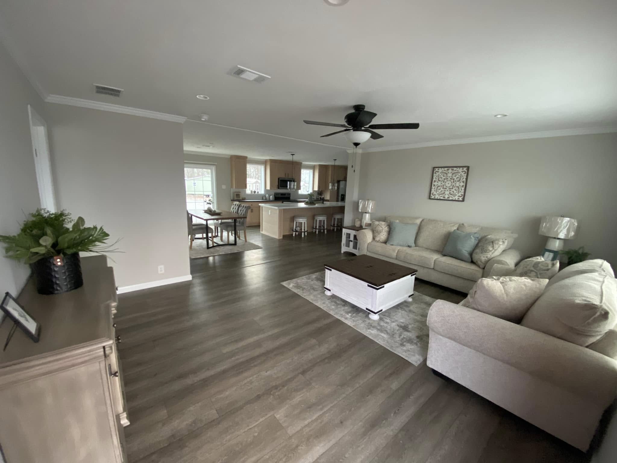 Spacious living room with light gray walls, wood flooring, beige sofas, and a ceiling fan. Open kitchen with a central island and dining area visible. Cozy and modern.