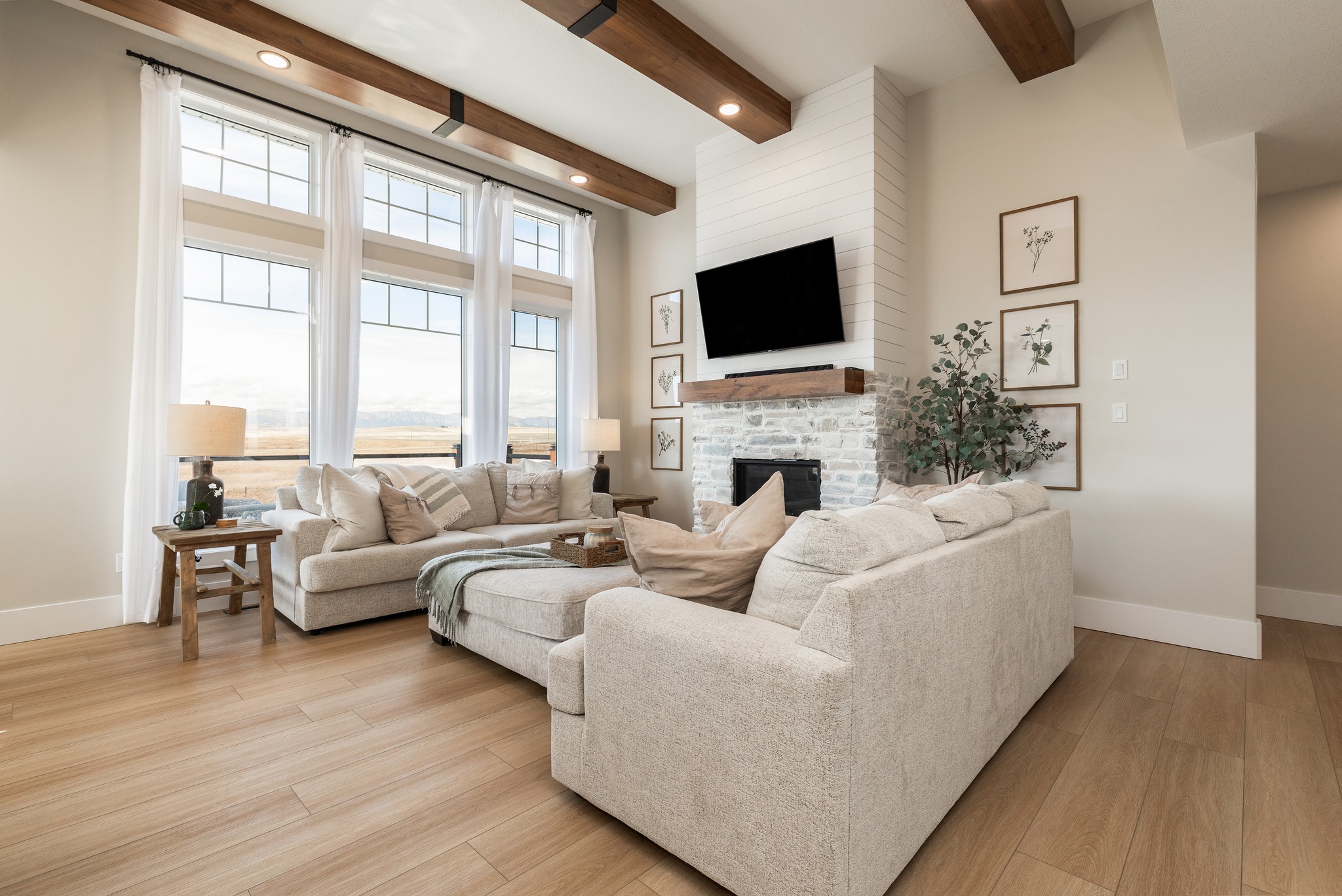 Cozy living room with beige sofas, large windows, and a brick fireplace. A TV is mounted above, with framed botanical prints on the walls. Bright and inviting.
