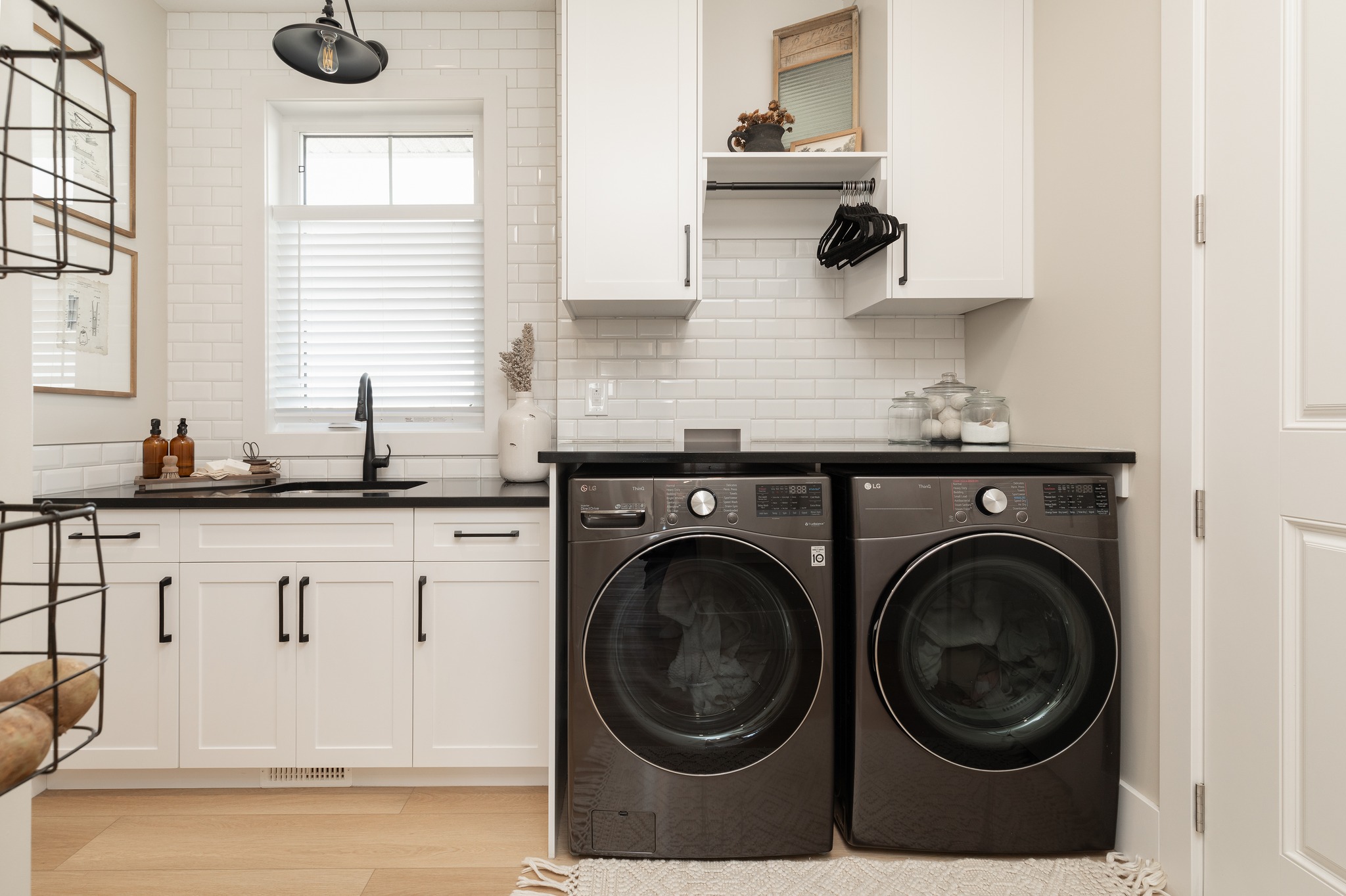 Modern laundry room with white cabinets, black countertops, and stainless steel appliances. A window with blinds and decorative jars add elegance.