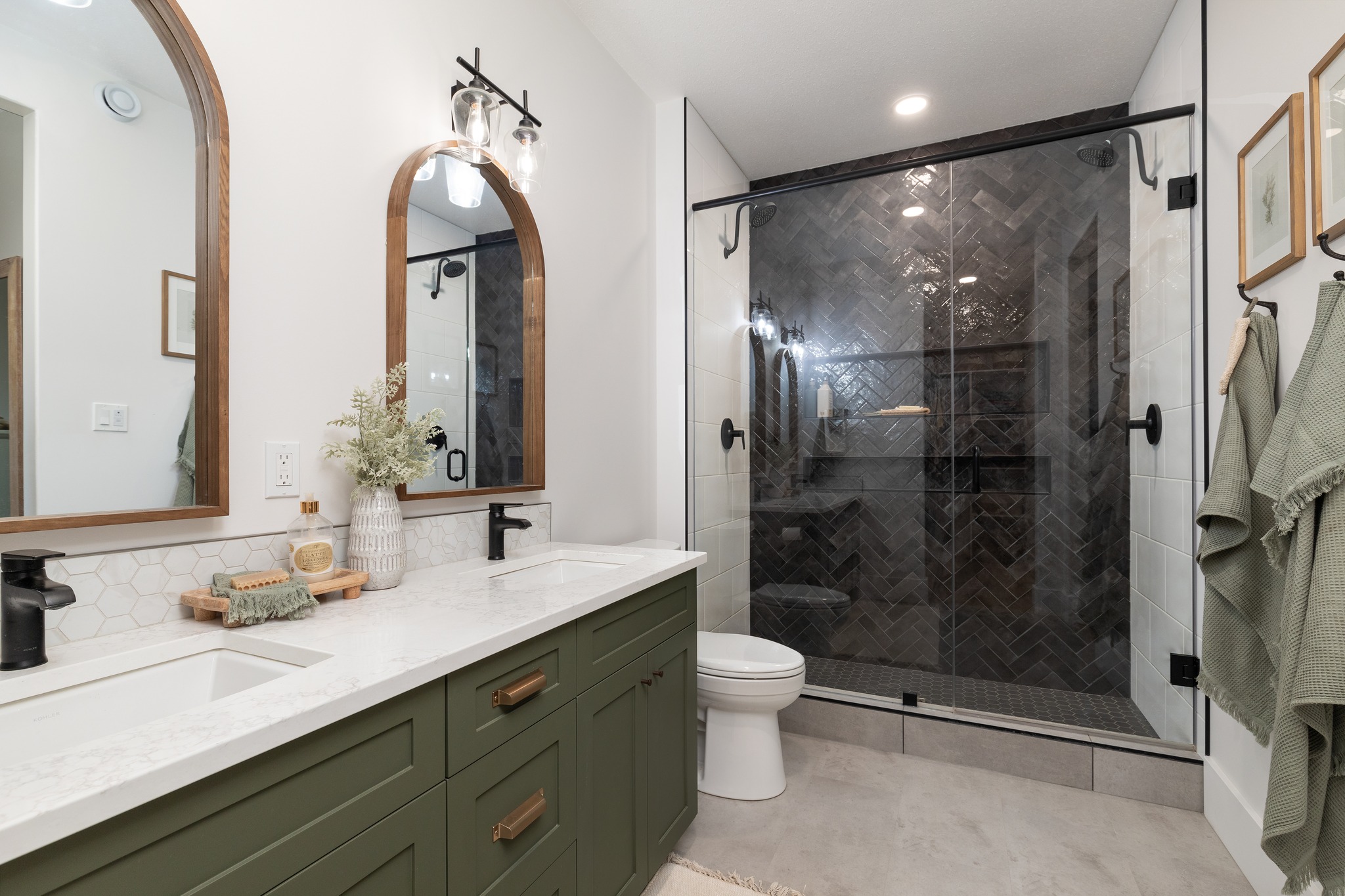 Modern bathroom with green vanity, white countertop, and dual arched mirrors. Features a glass-door shower with herringbone tiles and soft lighting.