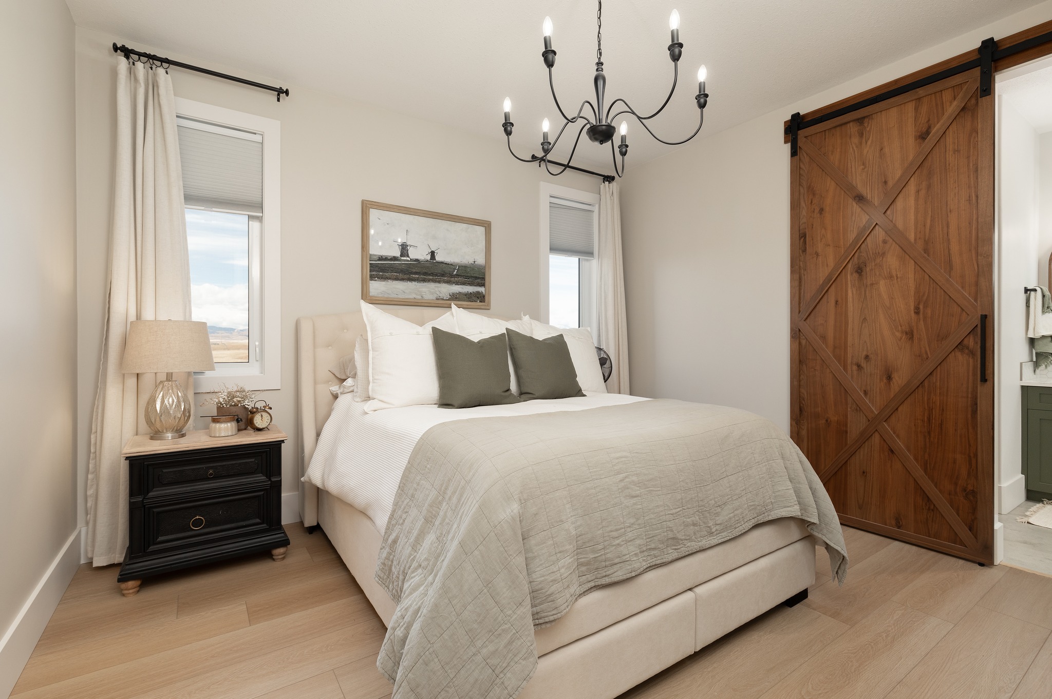 Cozy bedroom with a plush bed, white and green pillows, and a beige comforter. Features a rustic wooden sliding barn door, elegant black chandelier, and a landscape painting. Warm and inviting atmosphere.