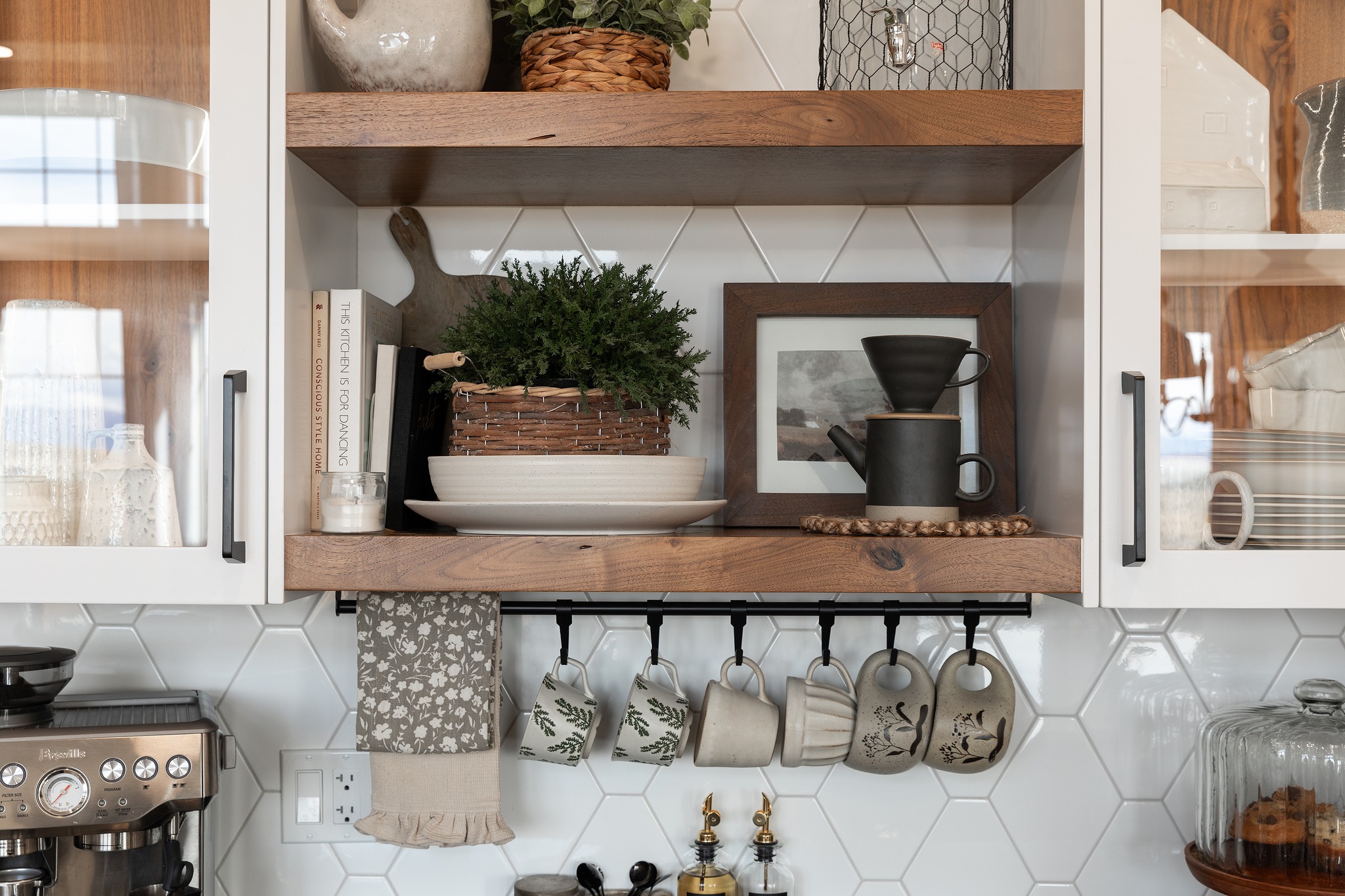 Warm, rustic kitchen shelf with wooden decor, potted plant, books, and framed art on hexagonal tile backsplash. Mugs hang below, creating a cozy ambiance.