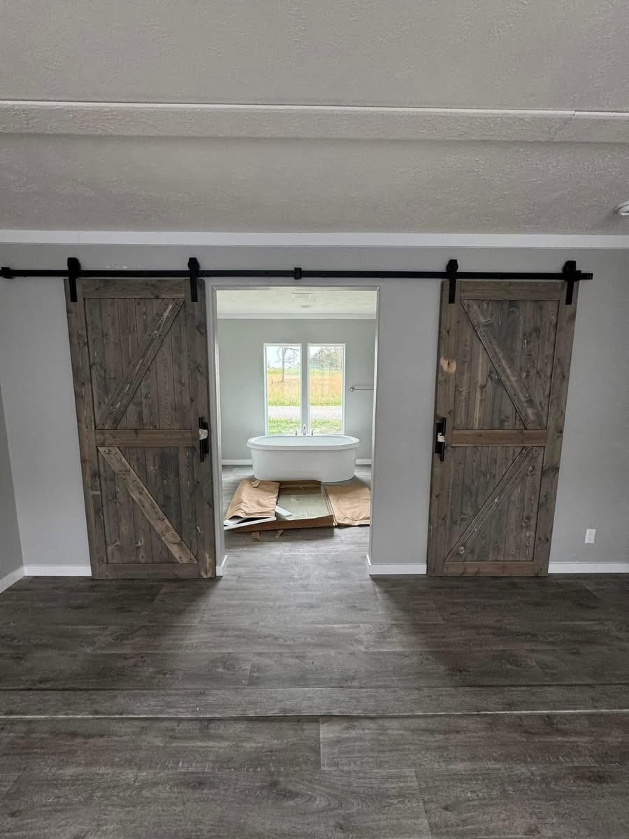 Gray room with rustic sliding barn doors open, revealing a bright bathroom with a white freestanding tub and large window. Calm and inviting atmosphere.