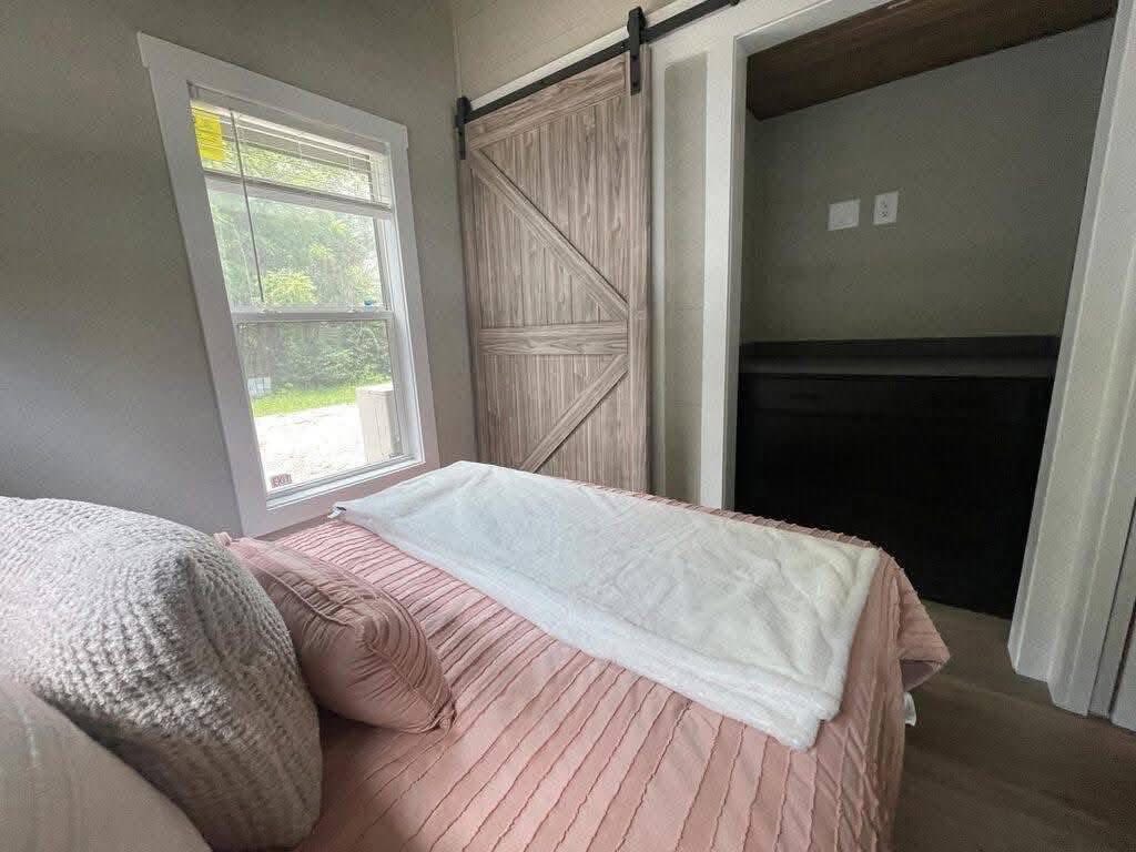 Cozy bedroom with a pink bedspread and pillows, beside a large window. A sliding barn door reveals a small closet, adding rustic charm.