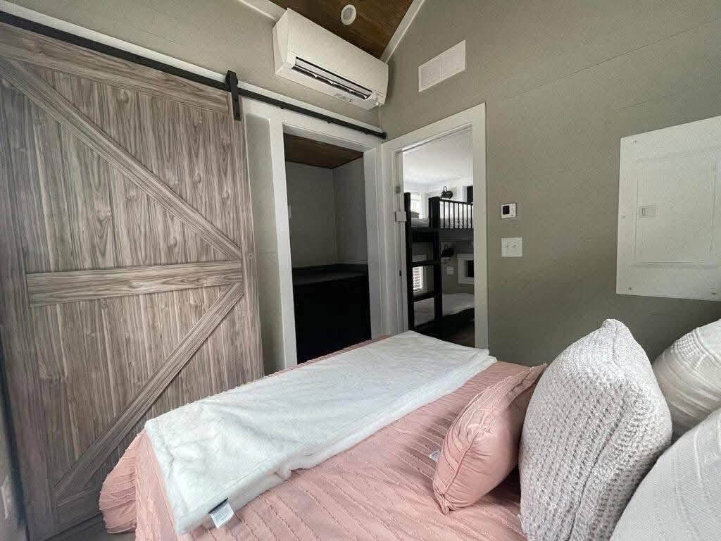 Cozy bedroom with a pink bedspread and soft pillows. A rustic wooden sliding door and modern air conditioning unit add warmth and comfort.