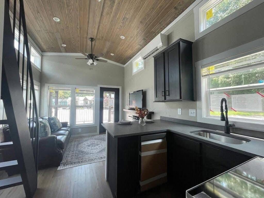 A modern tiny home interior features a wooden loft ceiling, sleek kitchen with black cabinets, and a cozy living area with a dark sofa and TV. Bright, airy feel.